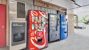 A row of vending machines, including an ICE dispenser and a Coca-Cola branded machine, outside a building with a wooden lattice design.