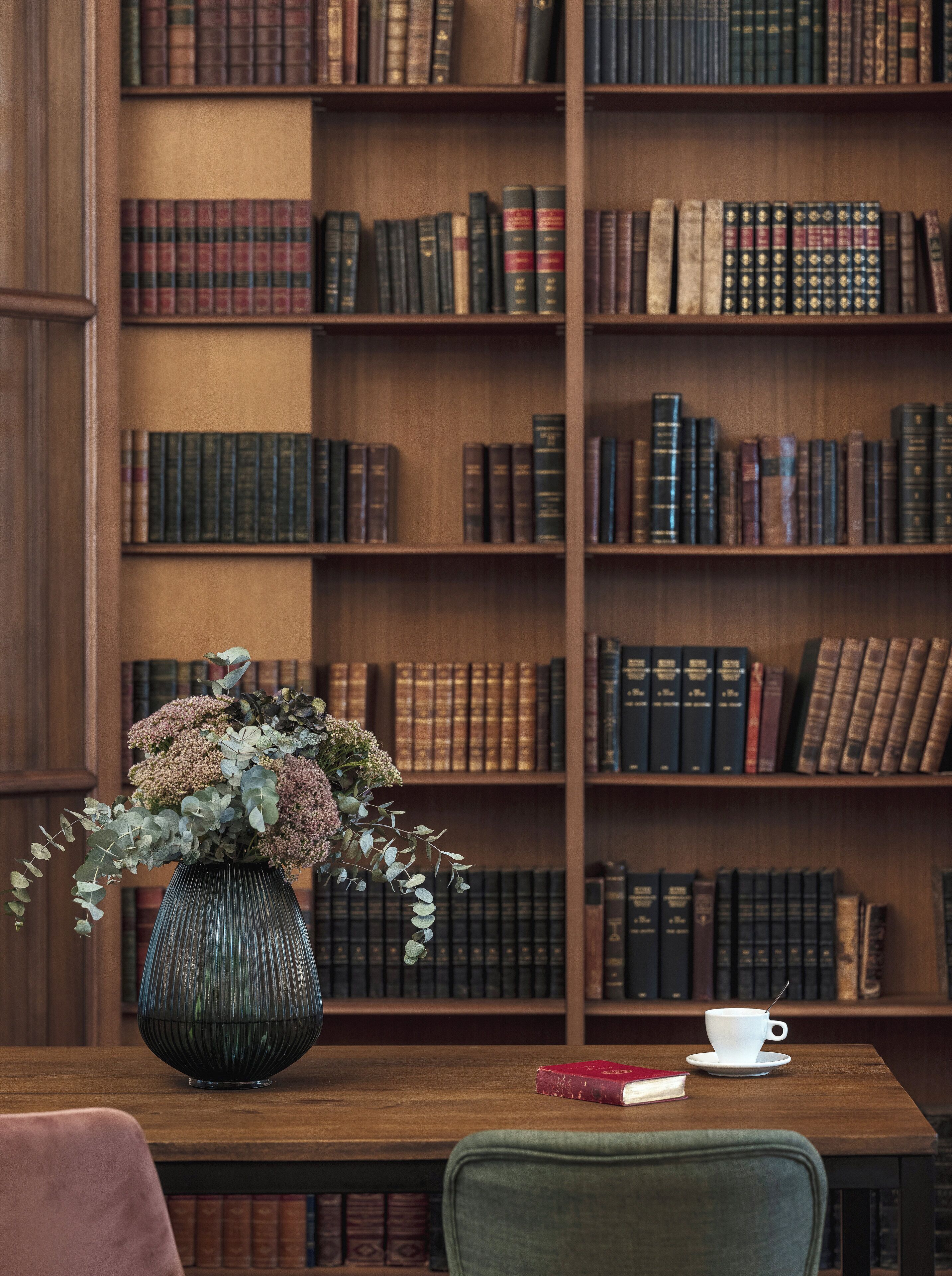 A wooden table with a vase of flowers, a book, and a cup. Behind the table is a bookshelf filled with books.
