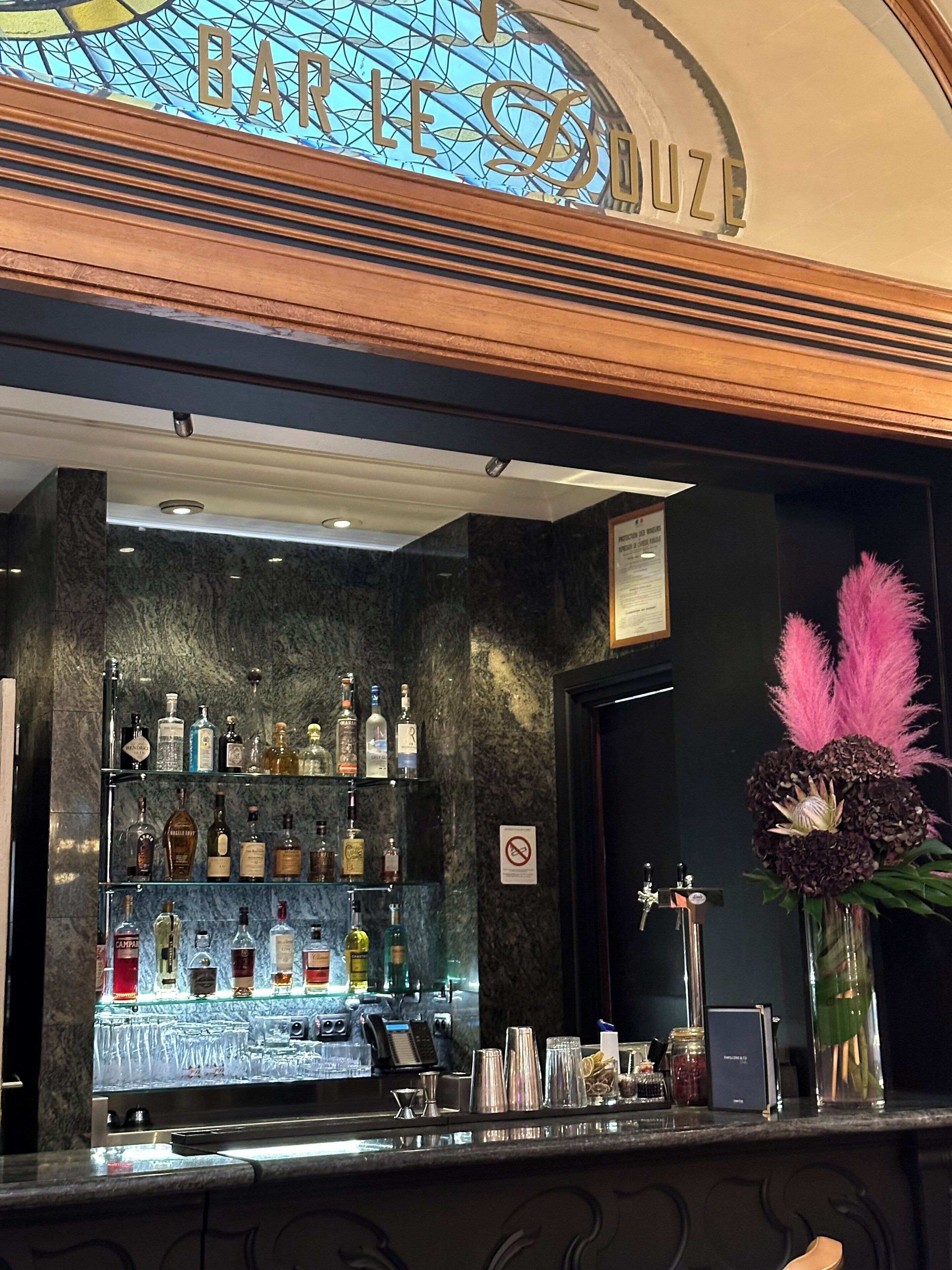Zona bar con scaffali di bottiglie di liquore, un bancone e un'elegante composizione floreale rosa.