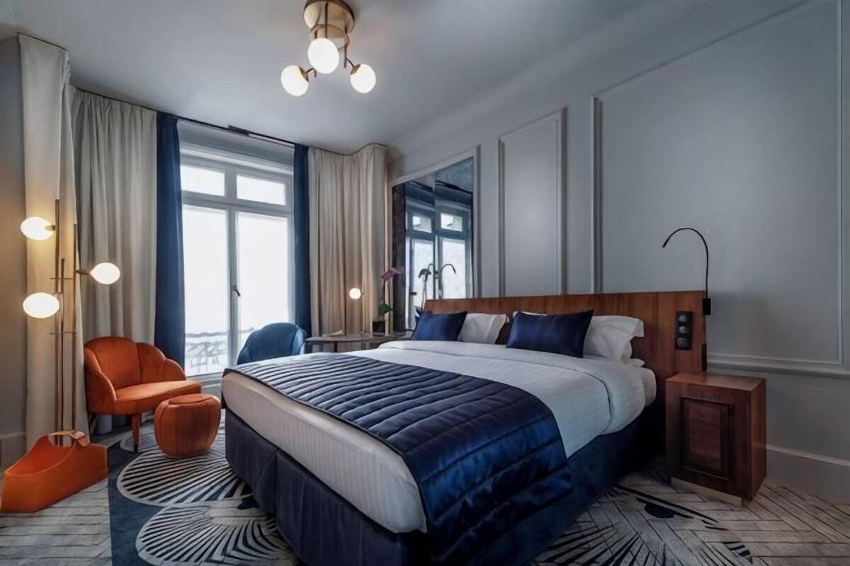 A modern hotel room with a large bed, a seating area with a chair, a bedside table, and a view of the outdoors.