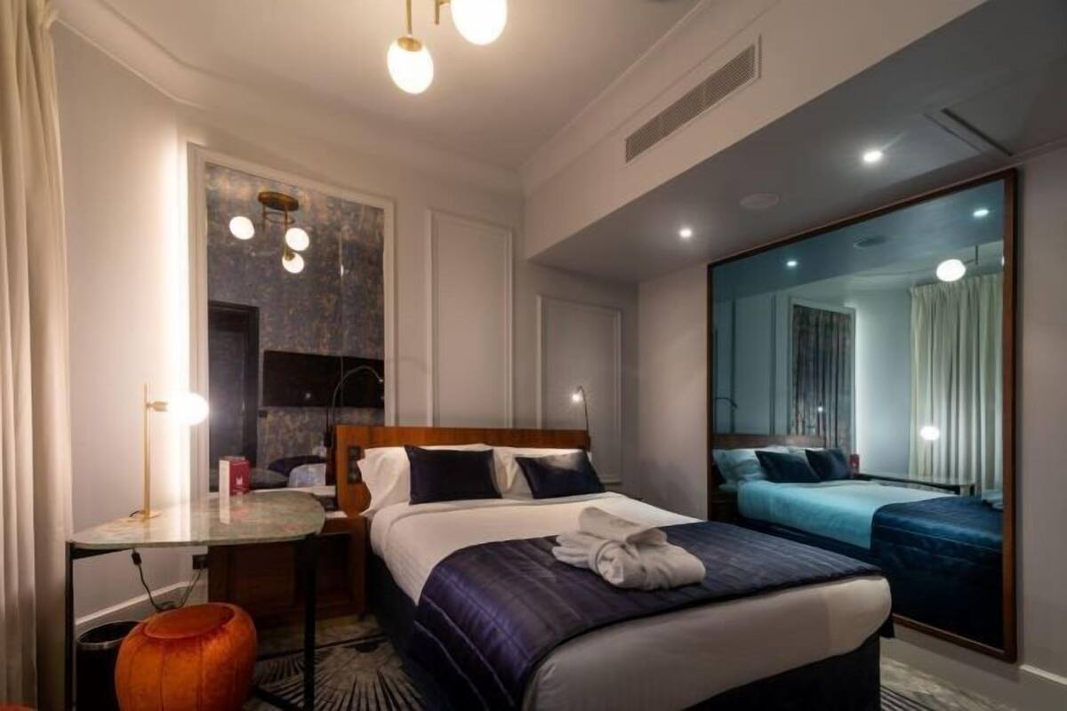A hotel room with a bed, bedside table, lamp, and a large mirror.