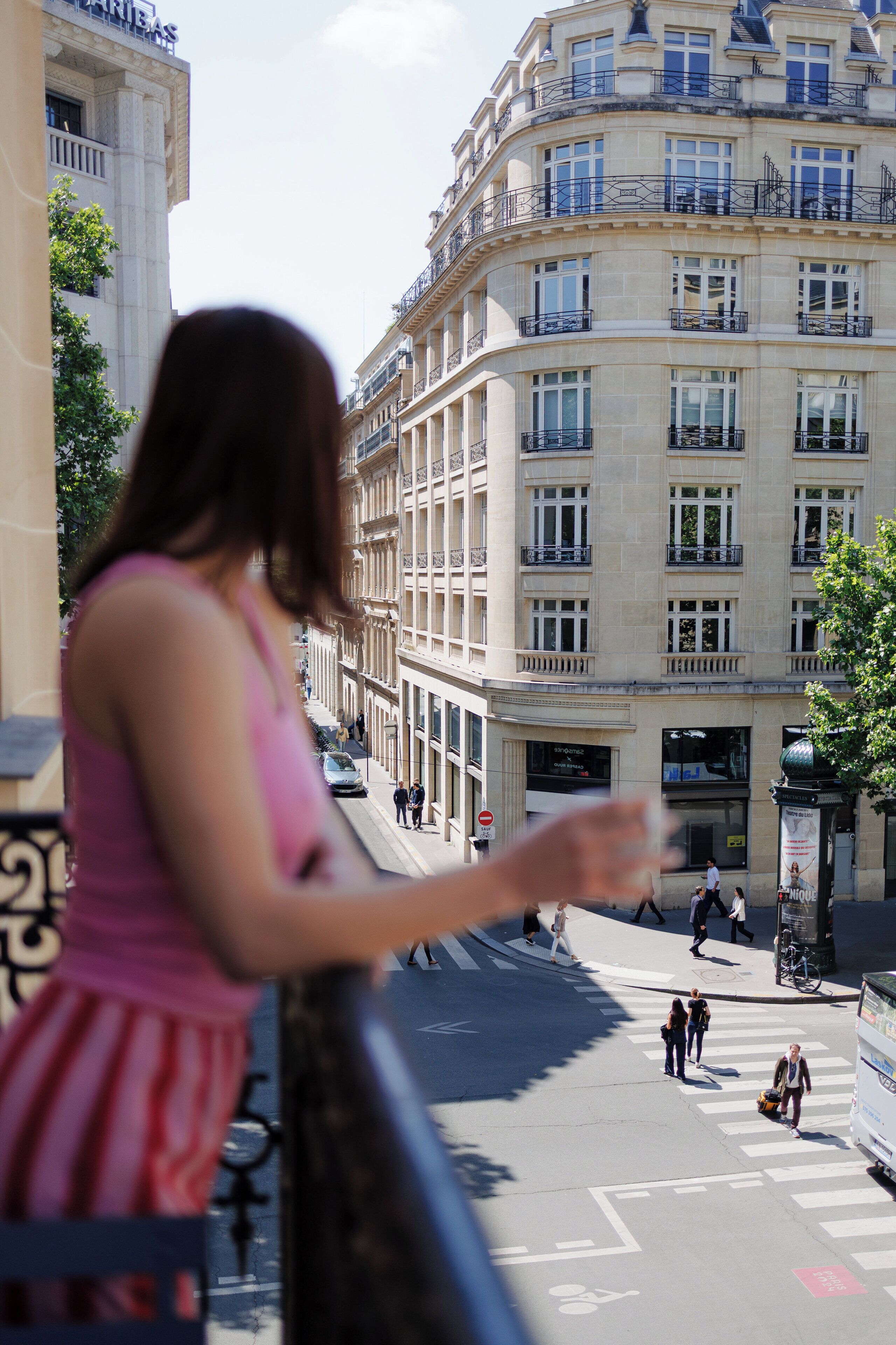 A woman standing on a balcony overlooking a street with buildings and pedestrians.