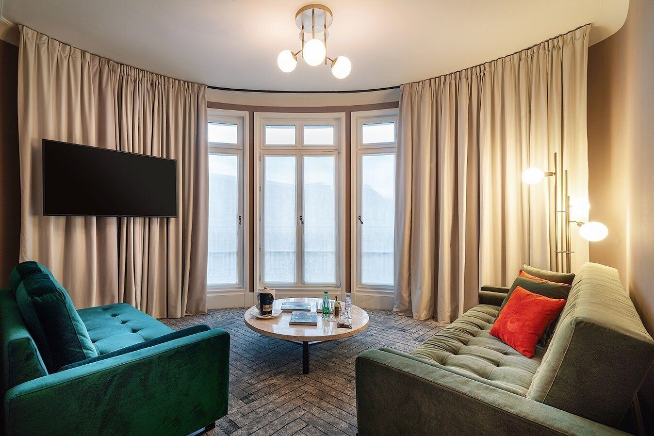 Un salon moderne avec un téléviseur à écran plat, deux fauteuils verts, un canapé vert avec des coussins rouges, une table basse ronde et une grande fenêtre avec des rideaux.