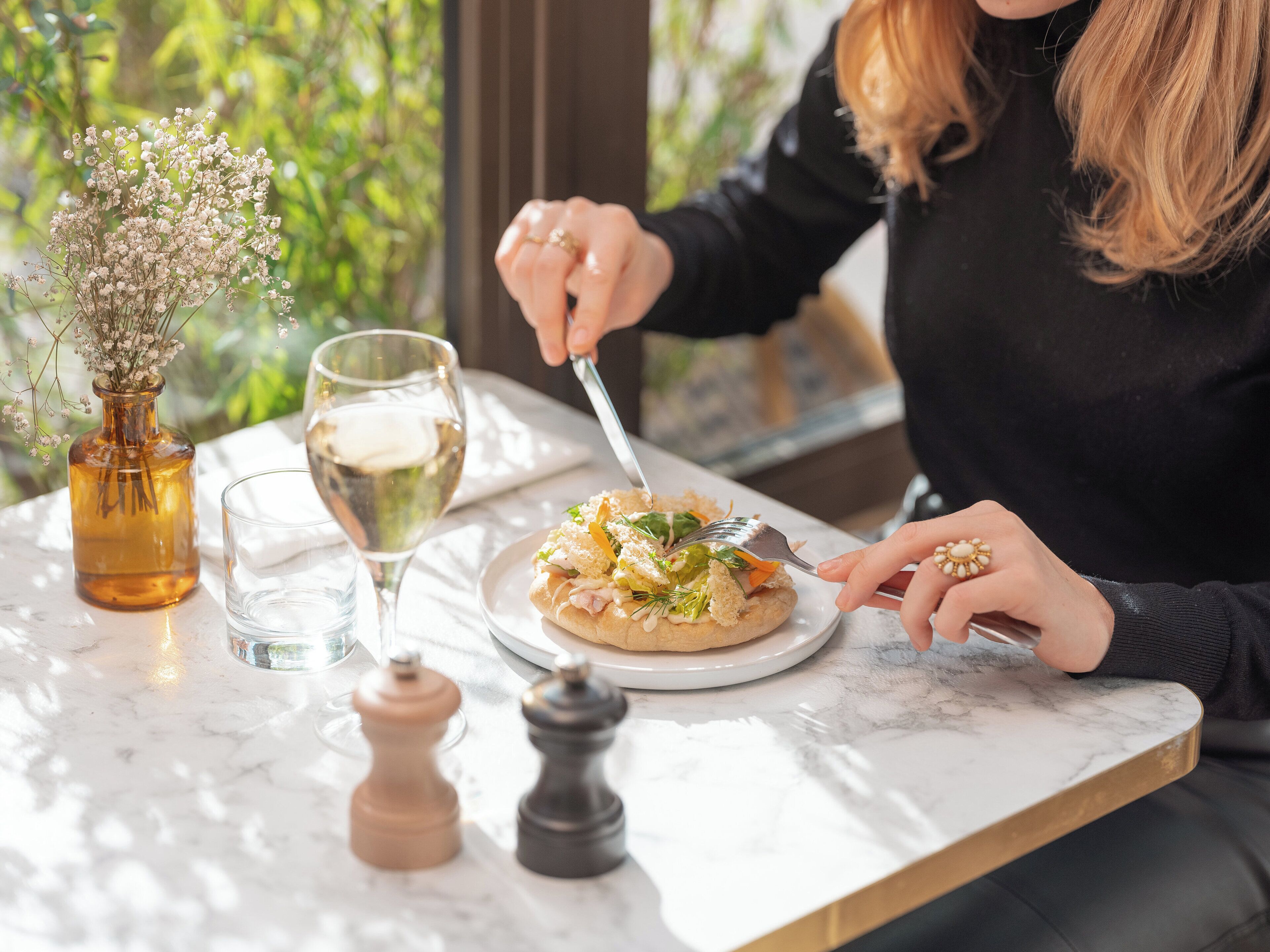 A person dining at a table with a plate of food, a glass of white wine, and a vase with flowers.
