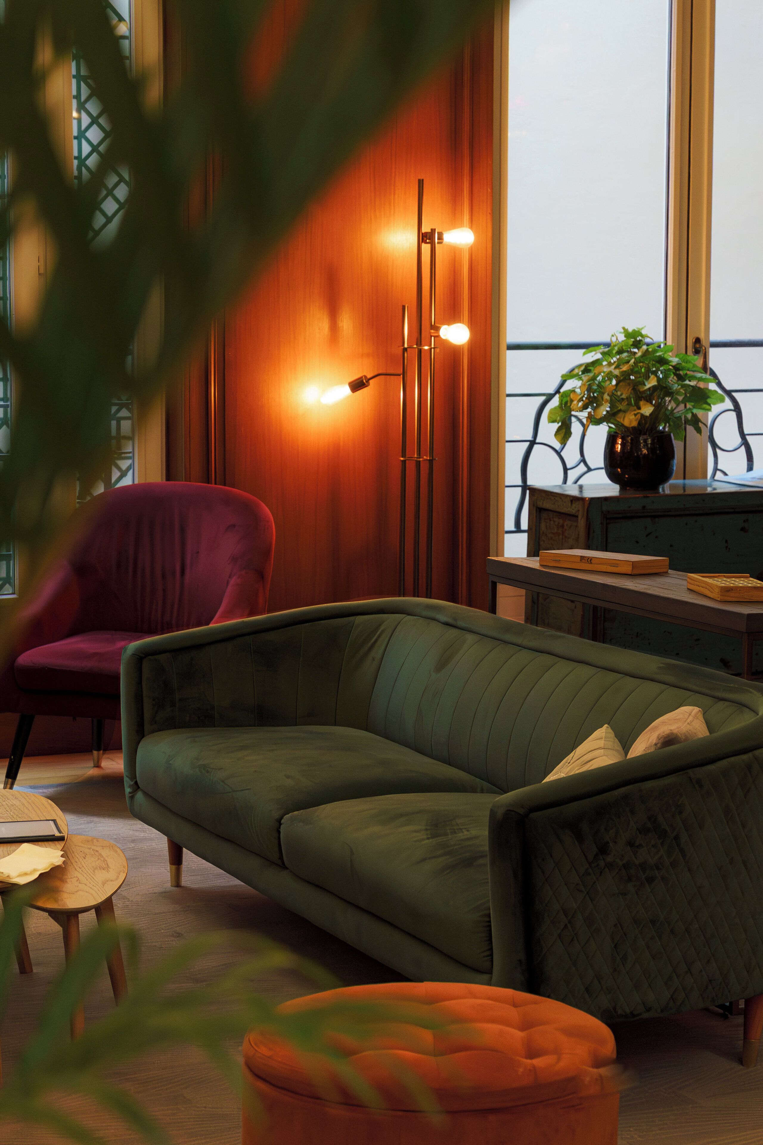 Una sala de estar acogedora con un sofá verde, un sillón rojo, una mesa de centro de madera y un pequeño otoman naranja. Hay una planta en maceta sobre un aparador azul y una lámpara de pie con tres luces.
