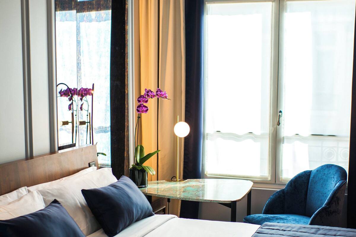 A hotel room with a large bed, a small table, a chair, and a window with curtains.