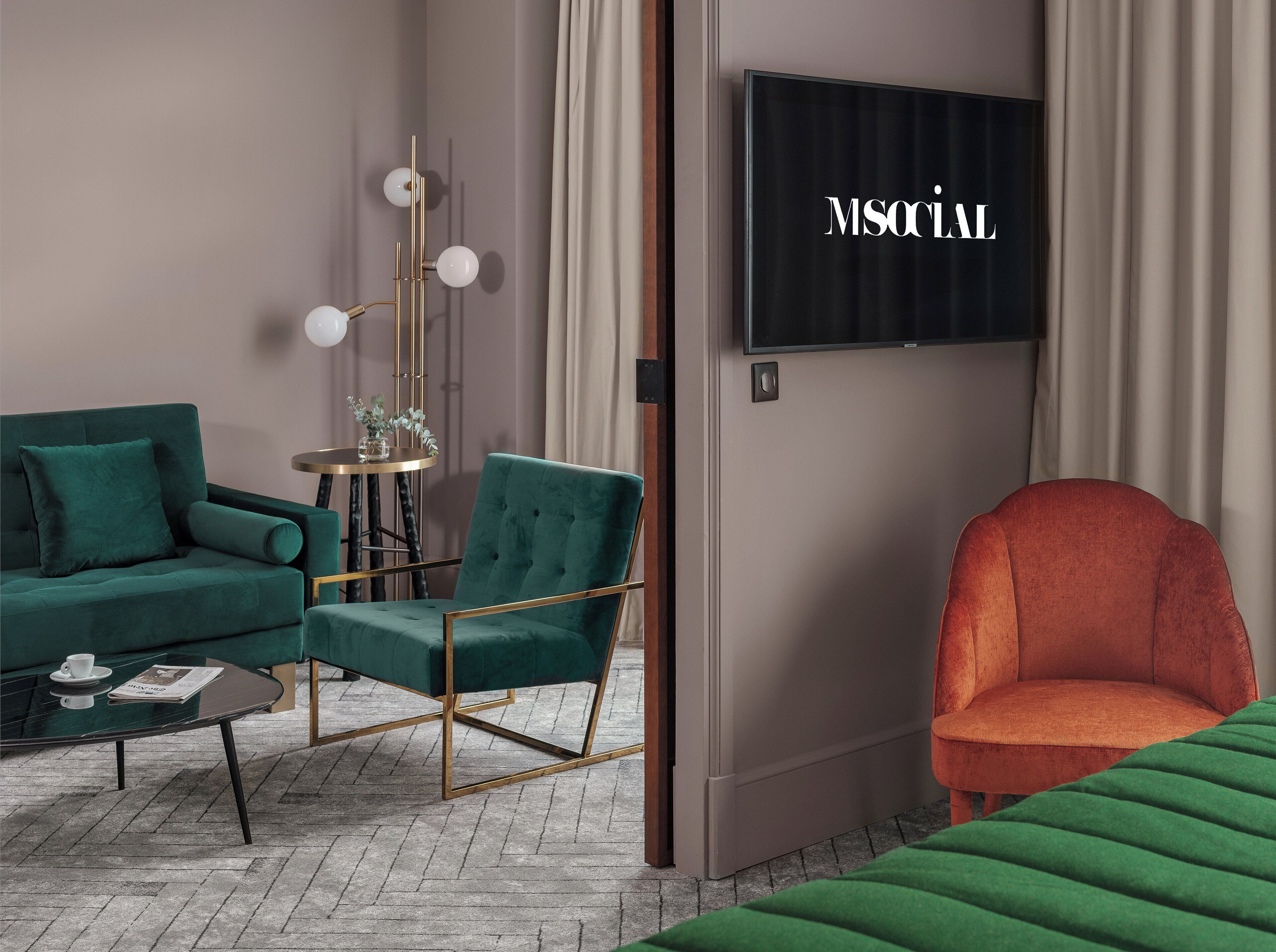 Una sala de estar moderna con un sofá verde, un sillón rojo y una mesa redonda con una lámpara. Un televisor de pantalla plana está montado en la pared.