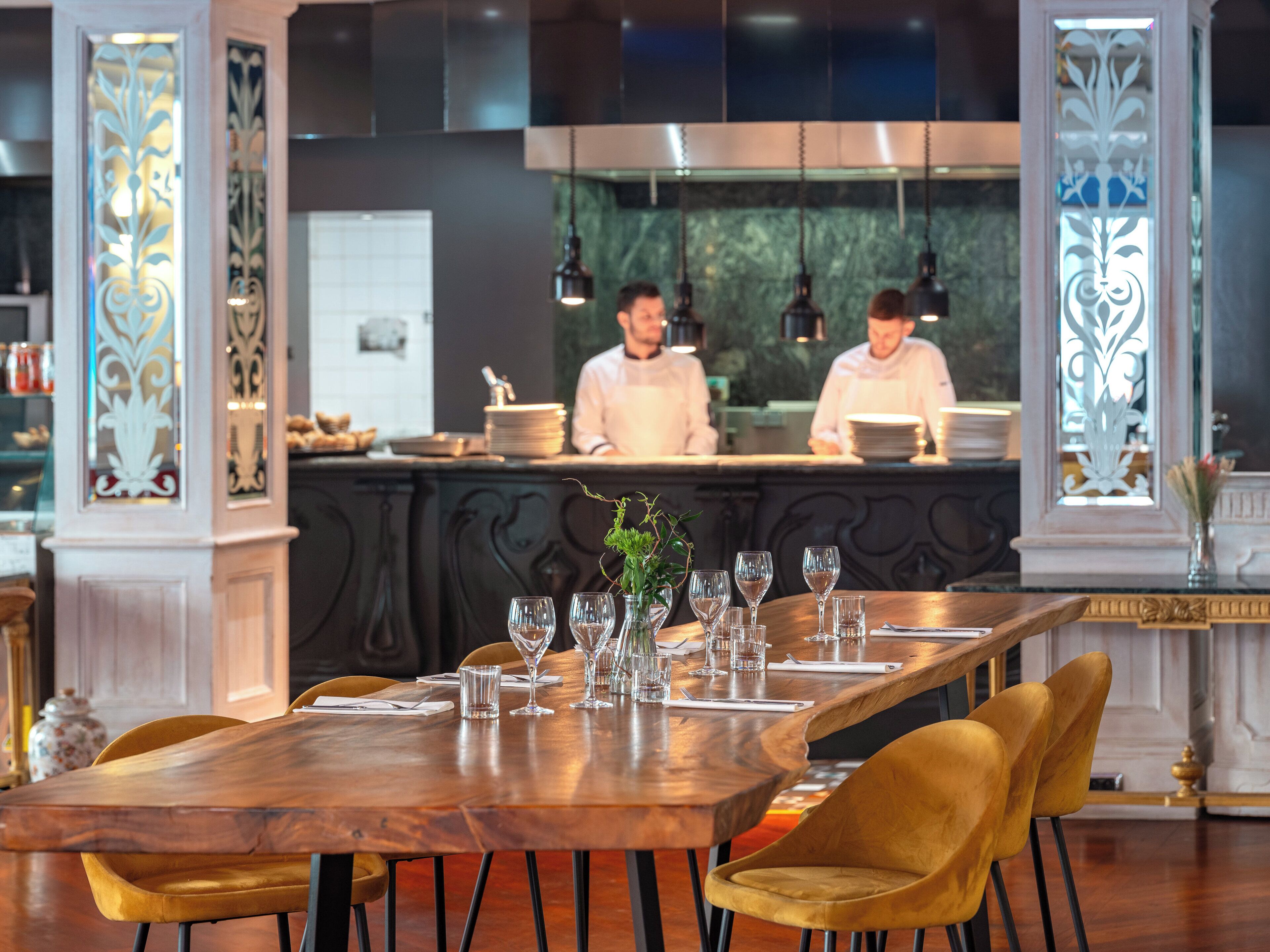 A restaurant interior with a long wooden table set for dining, gold chairs, and a bar area with chefs in the background.