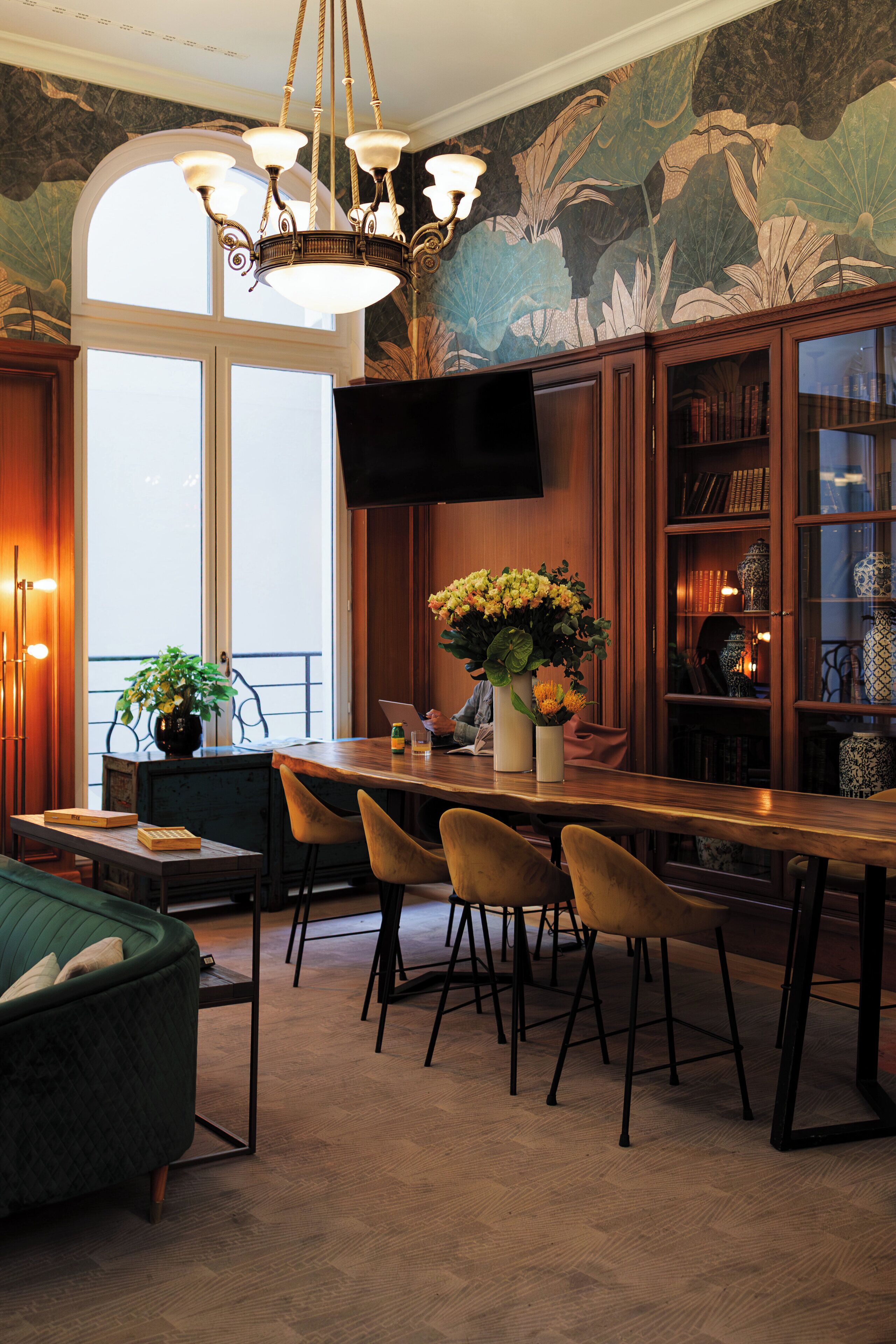 A dining area with a long wooden table, four chairs, a chandelier, and a large window with a view.