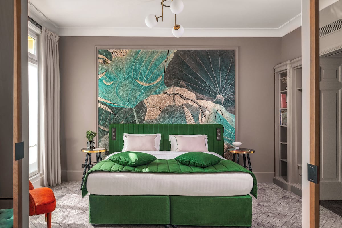 A bedroom with a green upholstered bed, a large abstract painting on the wall, and a modern chandelier.