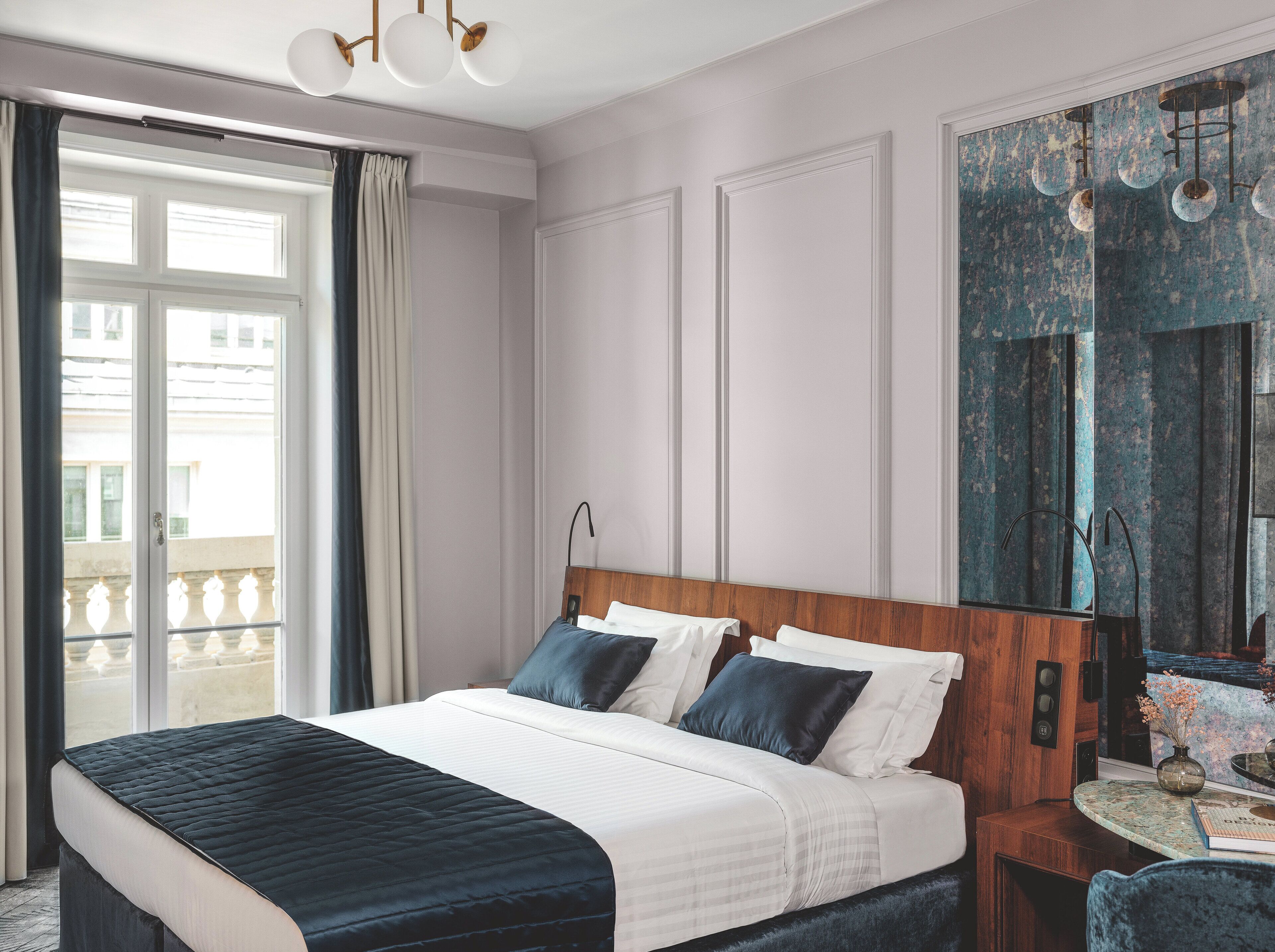 Una habitación de hotel con una cama grande, cabecera de madera, una mesa redonda con un florero y un espejo grande.