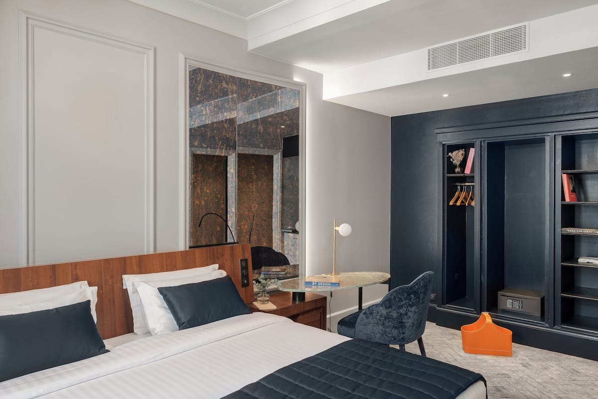 A modern hotel room with a large bed, a desk, a chair, a wardrobe, and a mirror.
