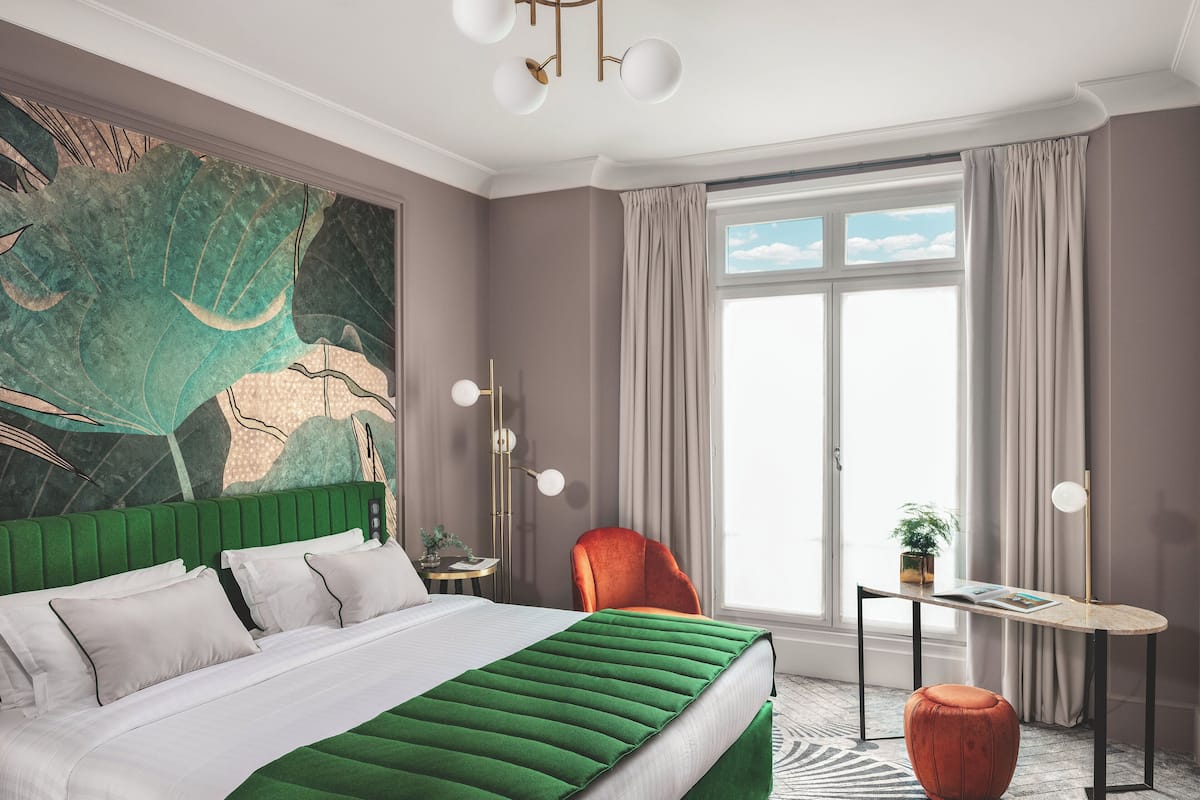 A modern bedroom with a large bed, a green accent wall, a view of the sky through a window, and a small table with a plant.
