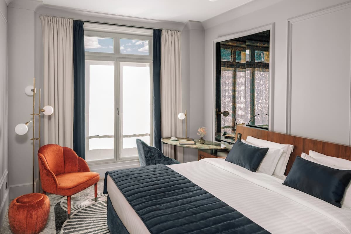 A hotel room with a large bed, a chair, a small table, and a view of the outdoors through a window.