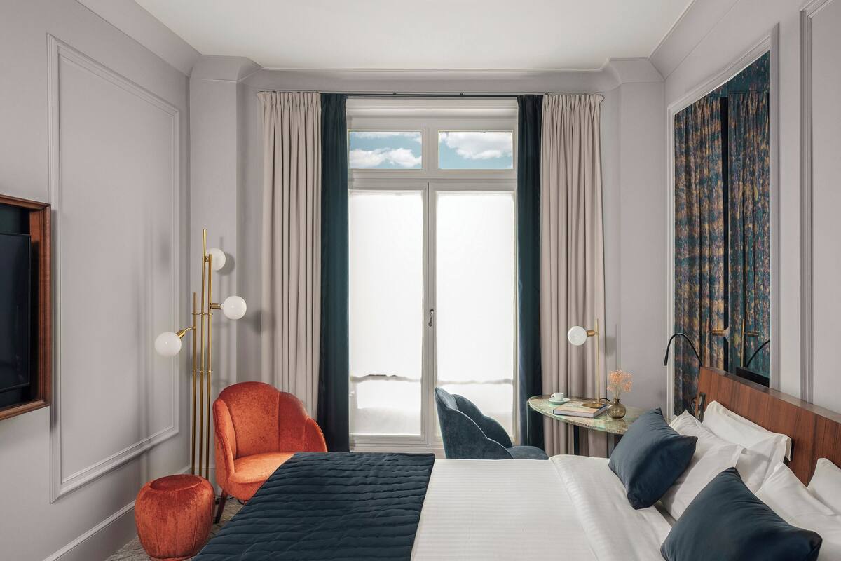 A hotel room with a large bed, a seating area with a chair and a small table, a television, and a window with curtains.