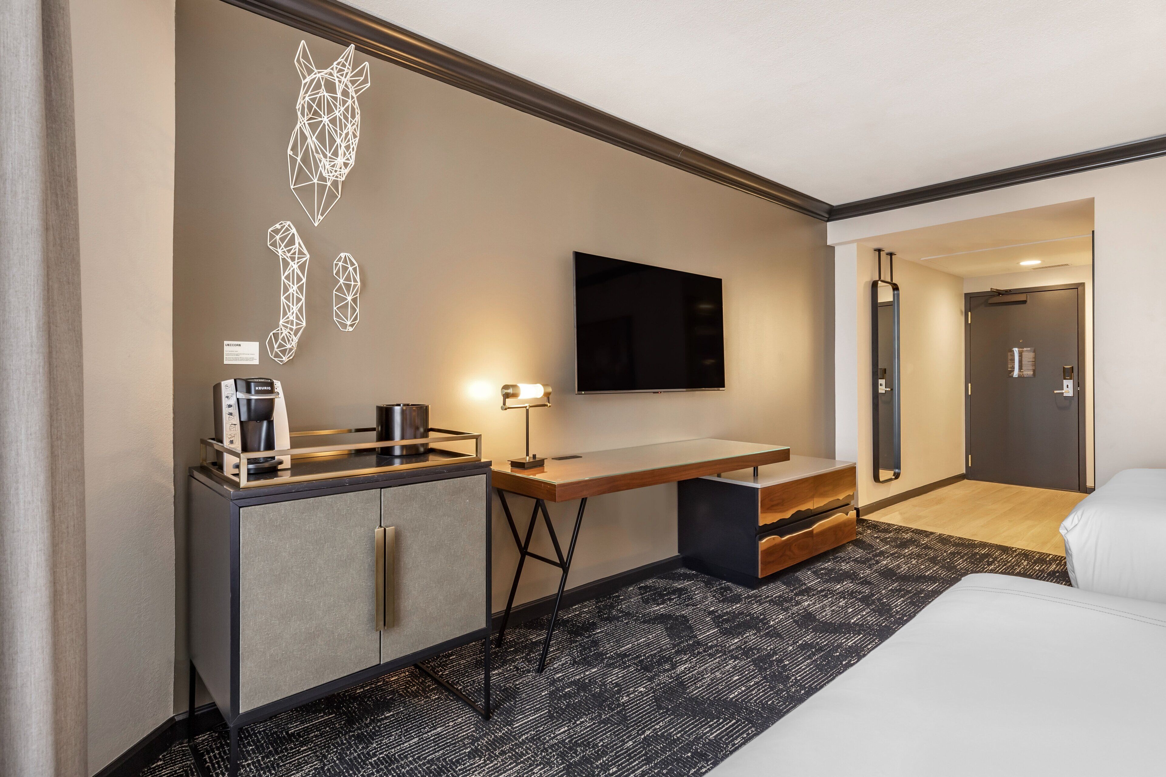 A modern hotel room with a bed, a desk, a coffee maker, a flat-screen TV, and a mirror.