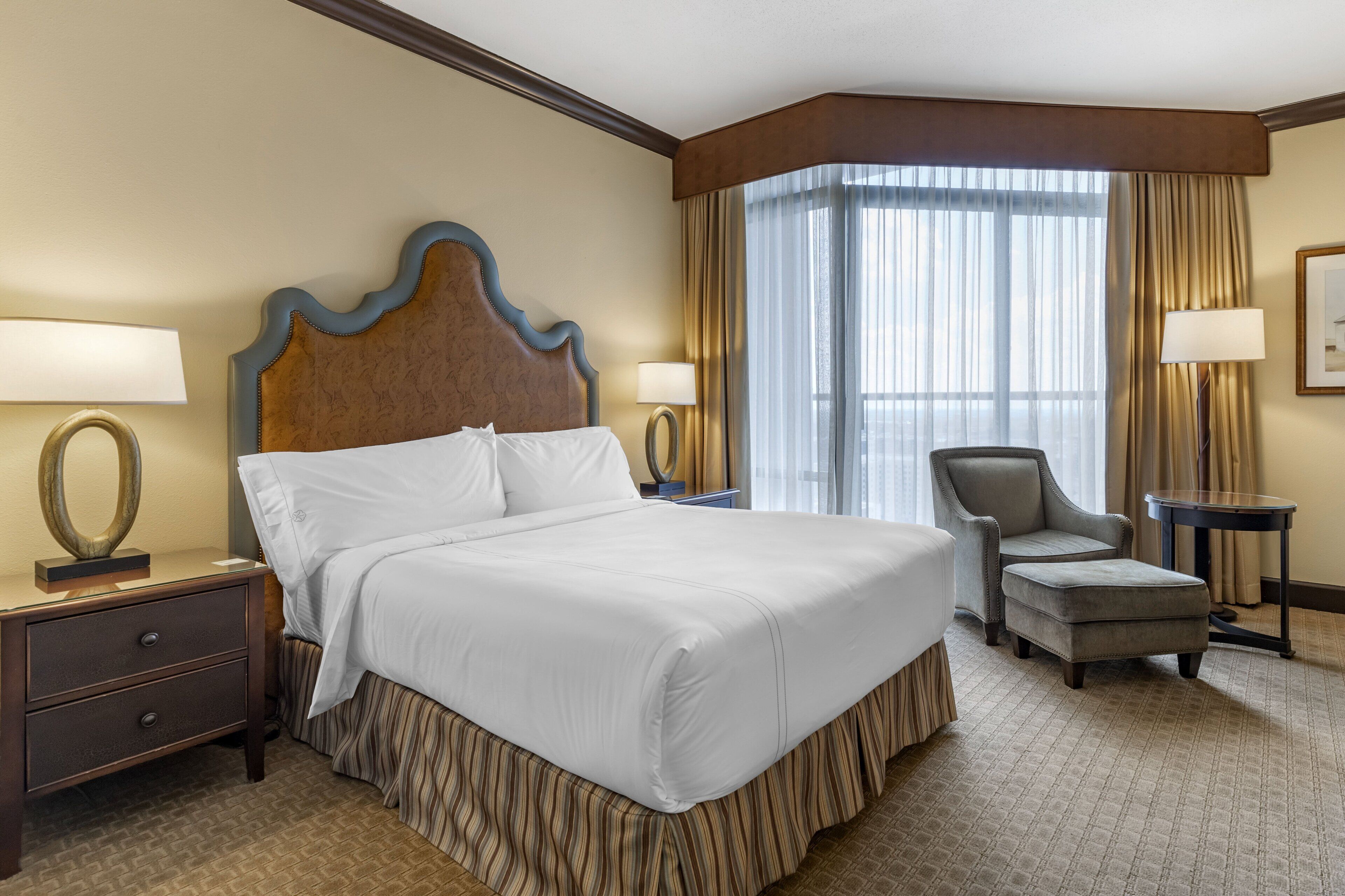 A hotel room with a large bed, two bedside tables, a chair, a desk, and a large window with curtains.