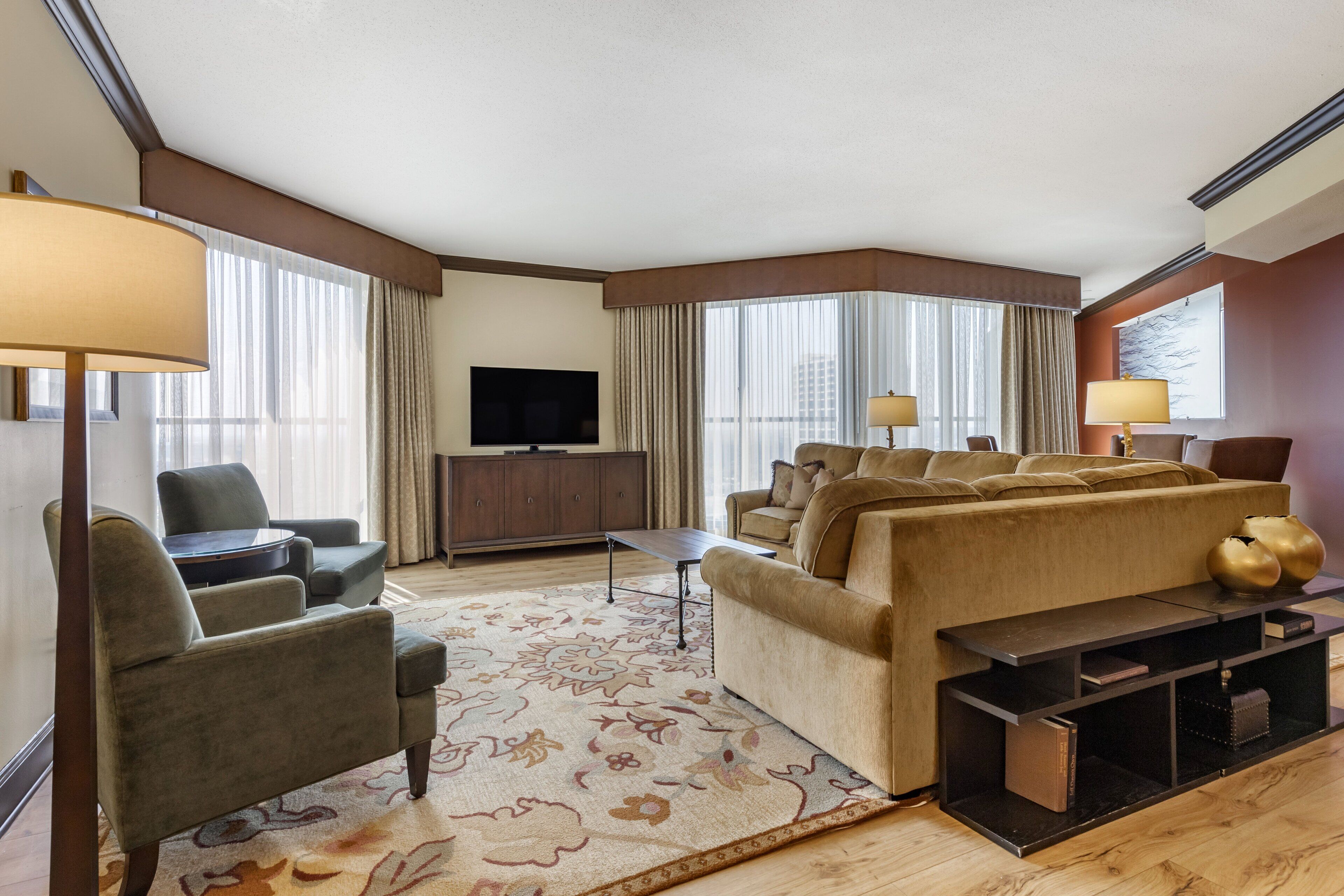 A modern hotel room with a large sofa, armchairs, a flat-screen TV, a coffee table, and a decorative rug.