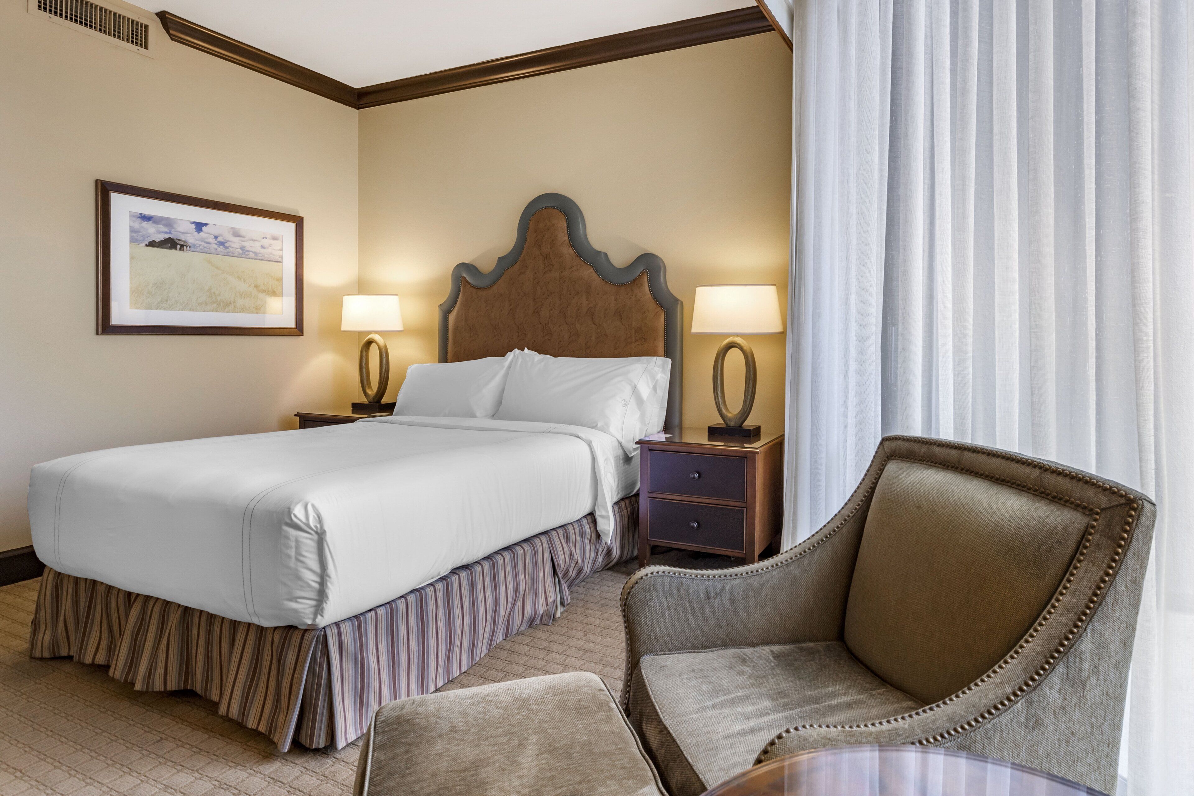 A hotel room with a large bed, two bedside tables with lamps, a chair, and a framed picture on the wall.