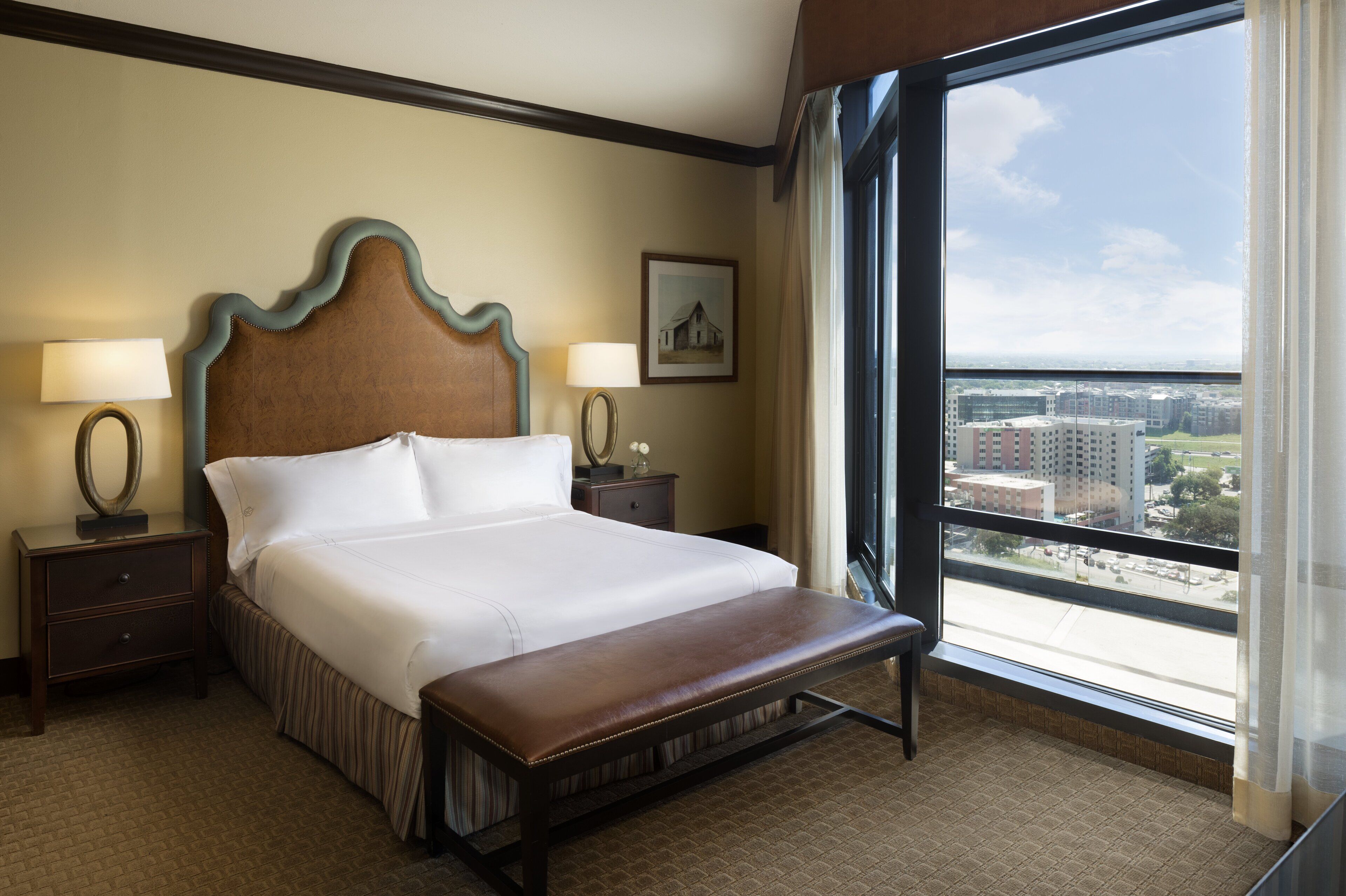 A hotel room with a large bed, two bedside tables with lamps, a bench at the foot of the bed, and a large window offering a city view.