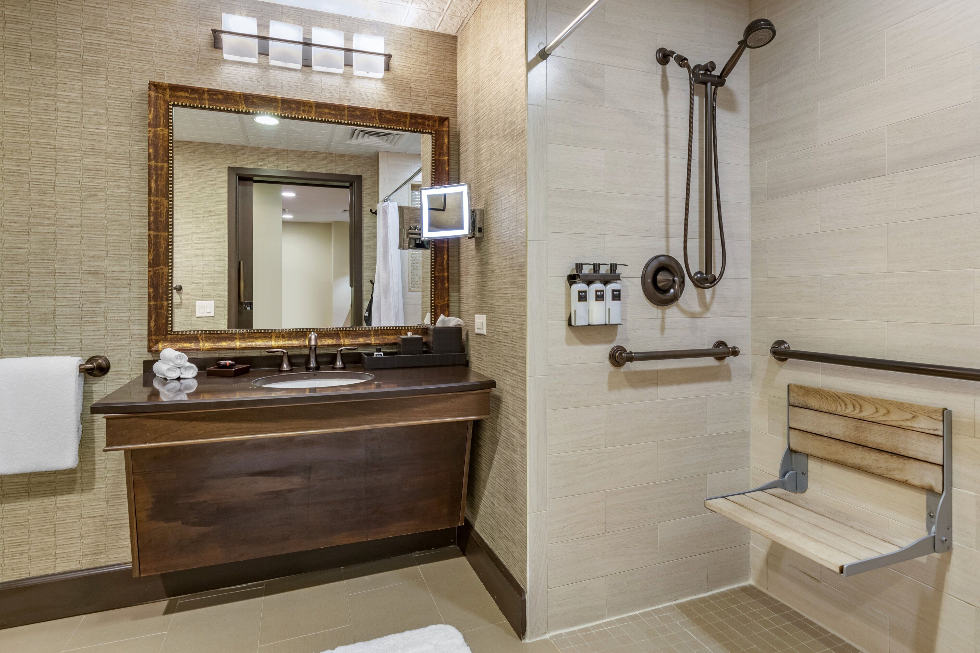Een moderne badkamer met een donker houten wastafelmeubel, een grote spiegel, een douche met handdouche en een houten bankje.