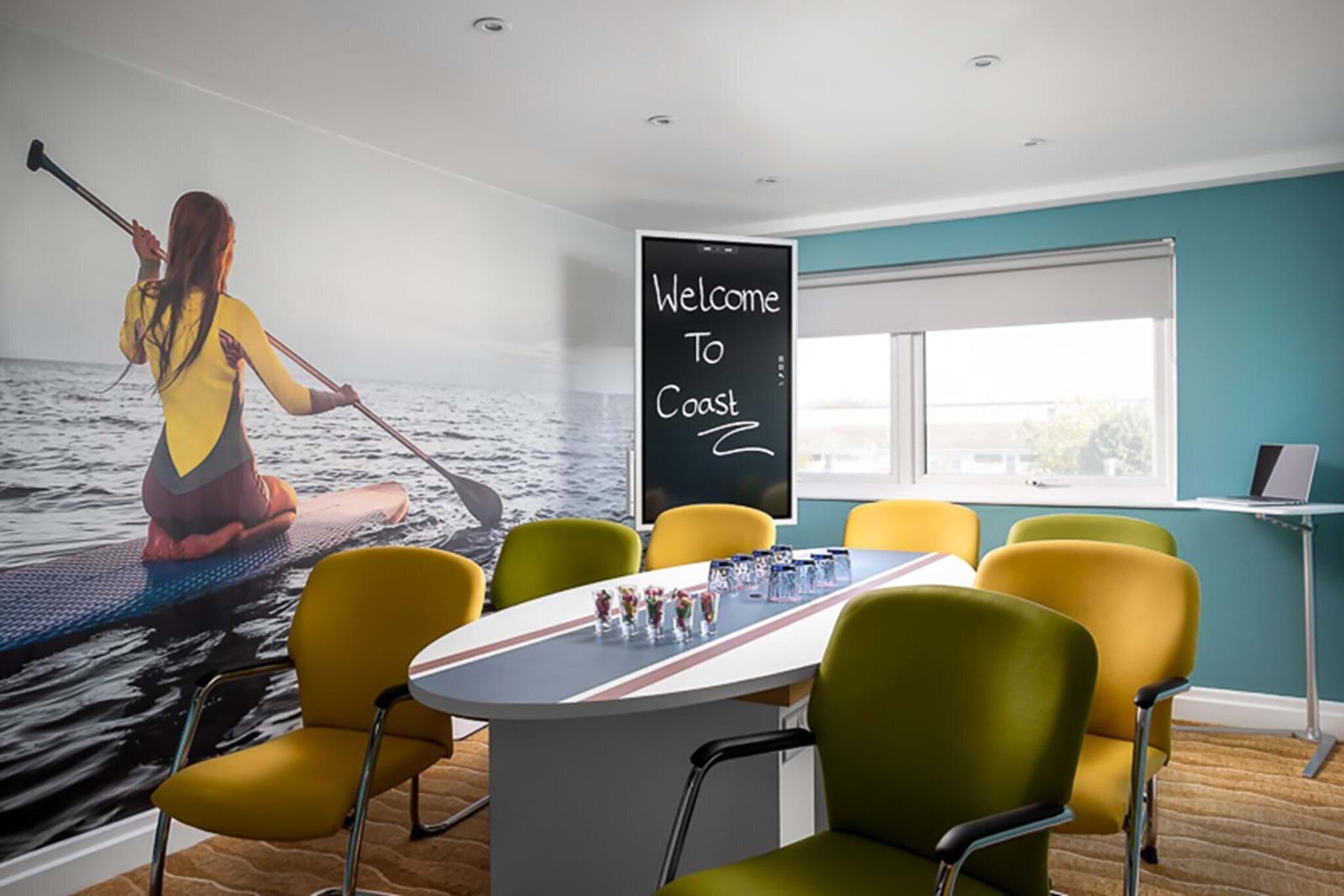 A room with a large window, a table with chairs, and a mural of a person paddleboarding. A sign on the wall reads "Welcome To Coast."