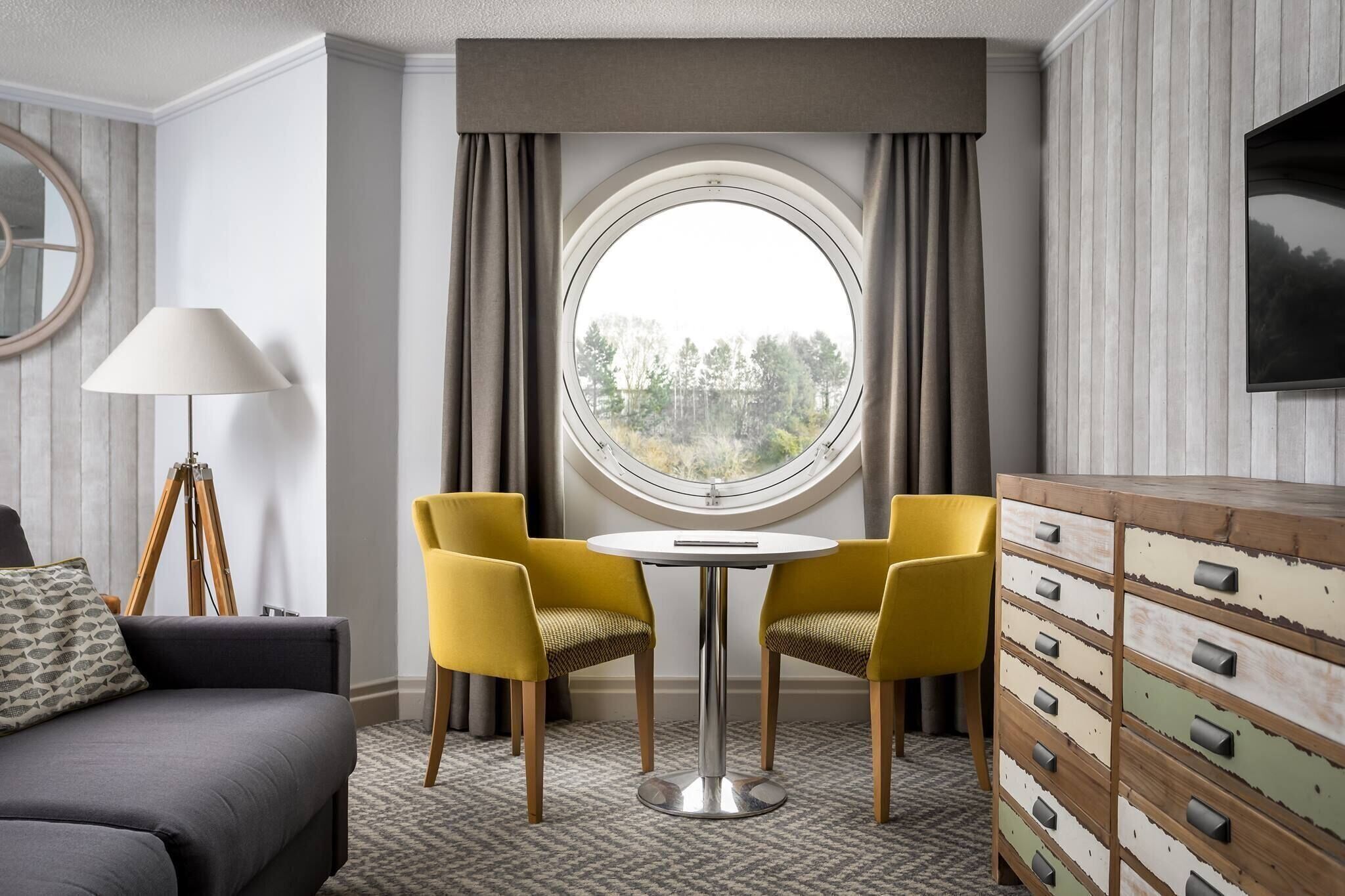Une pièce avec une fenêtre ronde, deux chaises jaunes, une petite table ronde, un canapé gris, une lampe et une commode en bois avec des tiroirs.