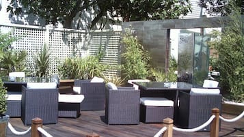 An outdoor seating area with wicker furniture and a wooden deck, surrounded by greenery and a lattice fence.