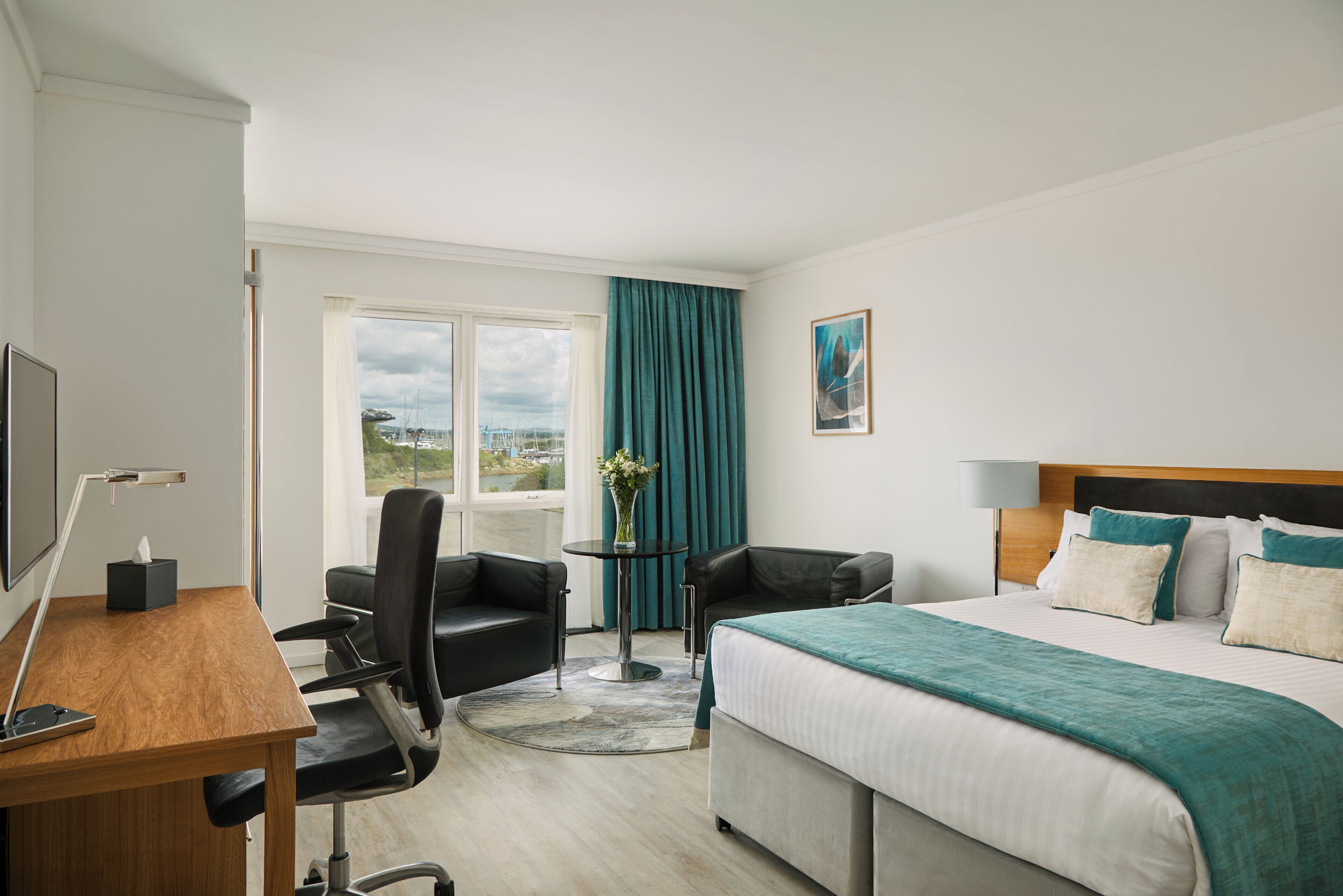 Ein Hotelzimmer mit Bett, Schreibtisch, Stuhl, Fernseher und einem Fenster mit Vorhängen sowie Blick ins Freie.