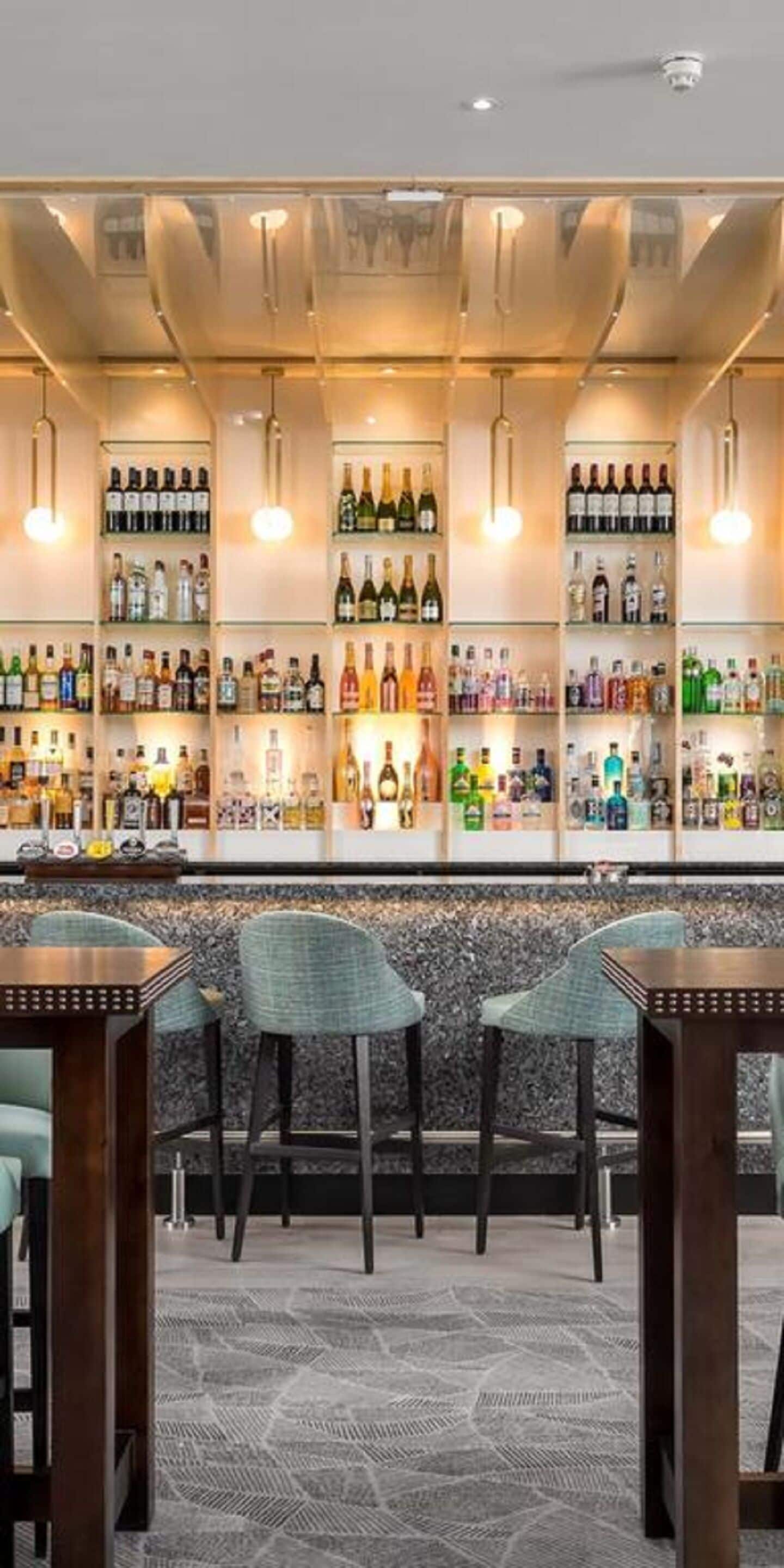 Zona de bar con varias estanterías llenas de botellas, una barra y taburetes.