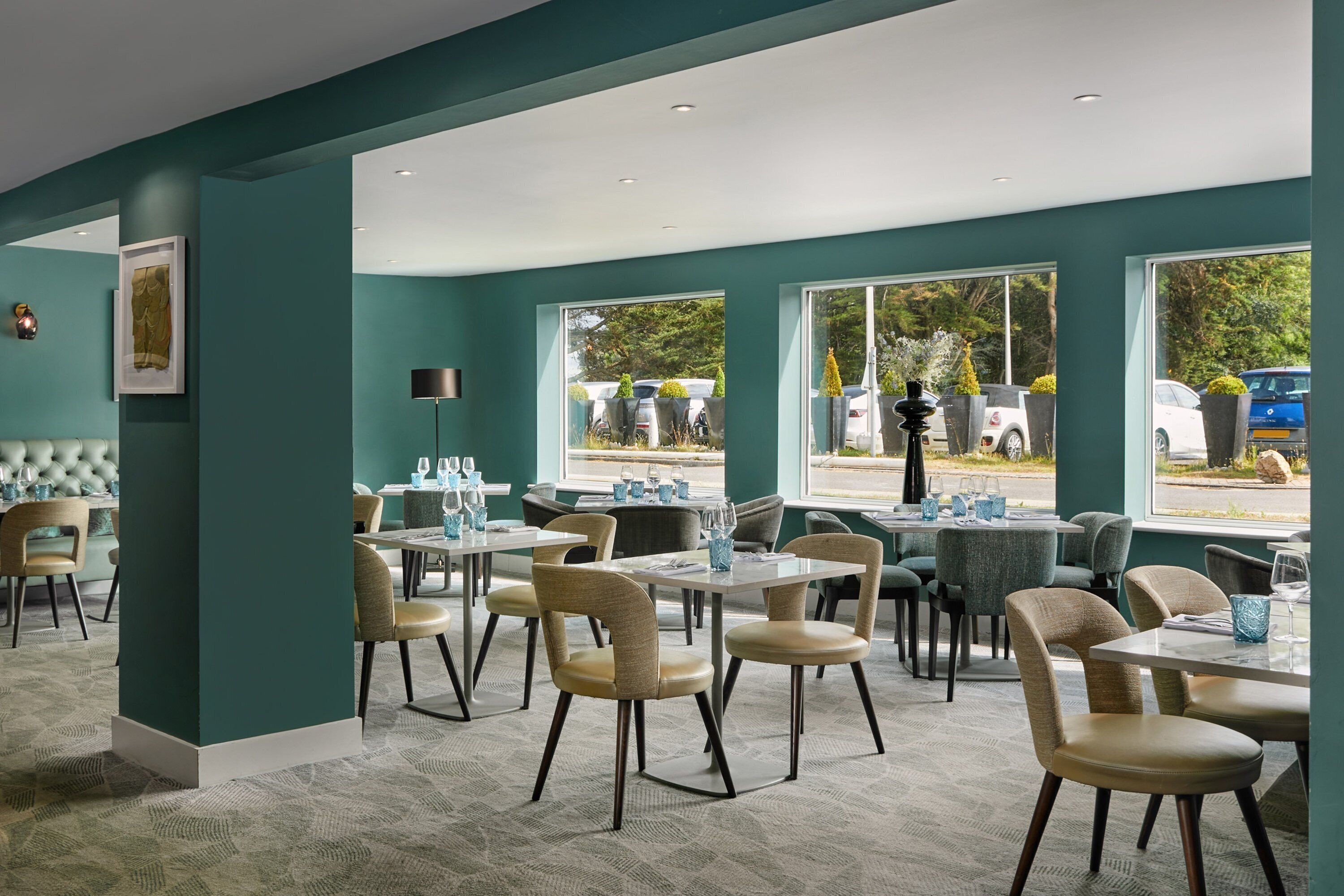 A modern dining area with teal walls, large windows, and a mix of round and rectangular tables.