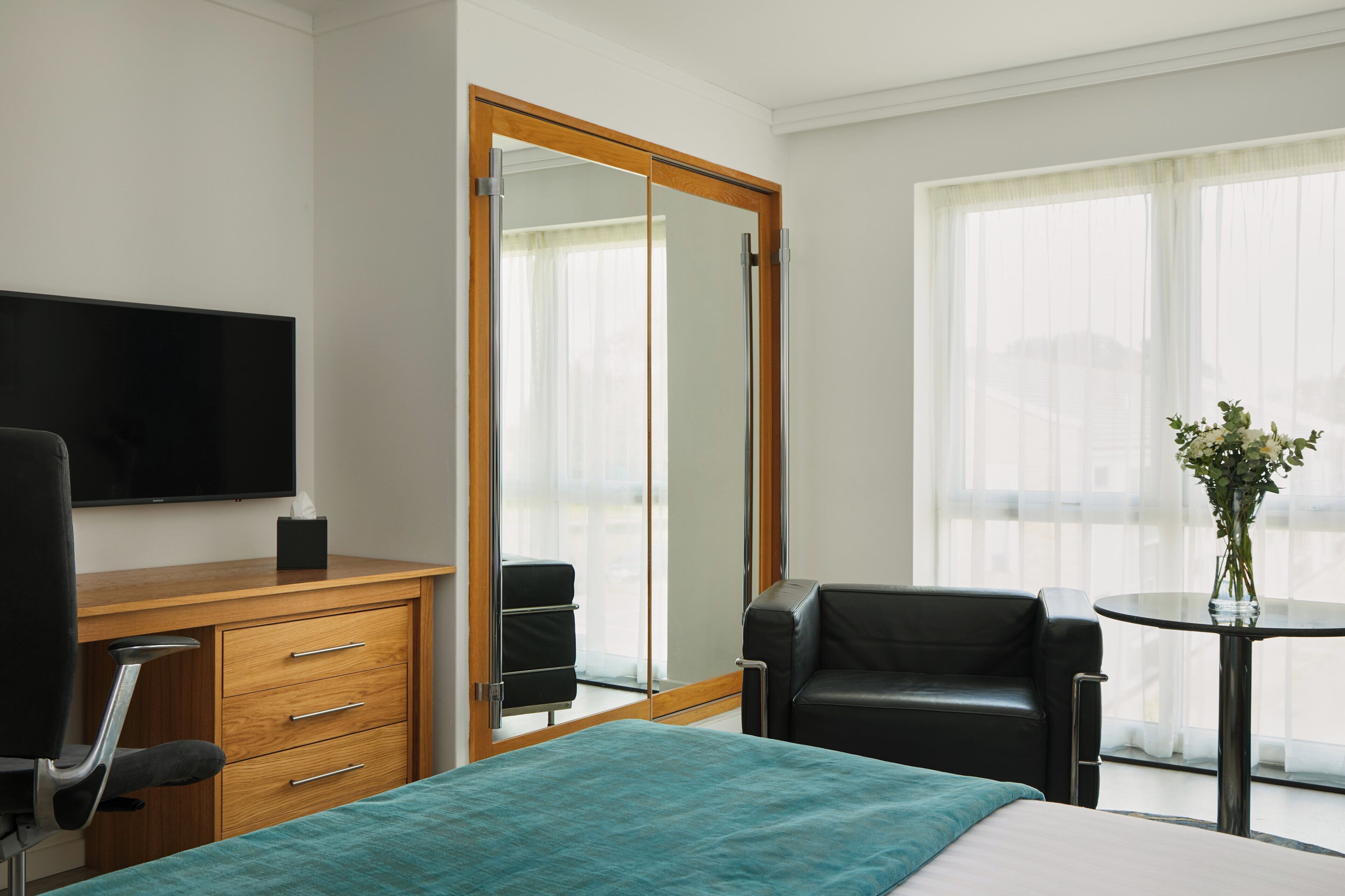 A hotel room with a bed, a wooden desk with a chair, a TV, a mirror, a black leather sofa, a small table with a vase, and large windows with sheer curtains.
