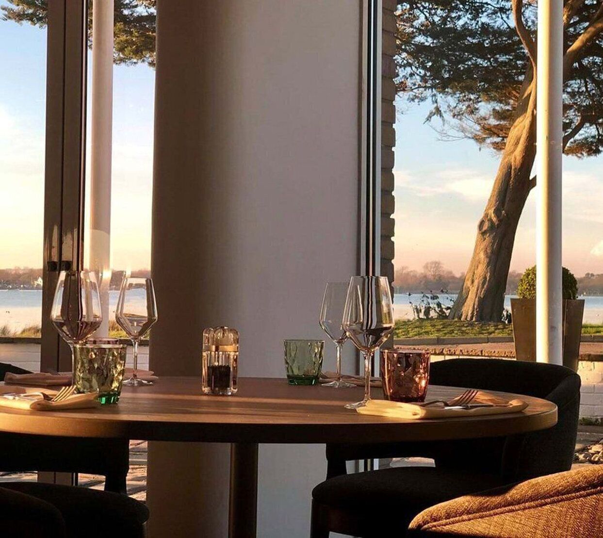 A dining table set with wine glasses and salt shakers, overlooking a lake with trees and a clear sky.
