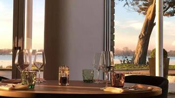 Mesa de comedor dispuesta con copas de vino y saleros, con vistas a un lago con árboles y cielo despejado.