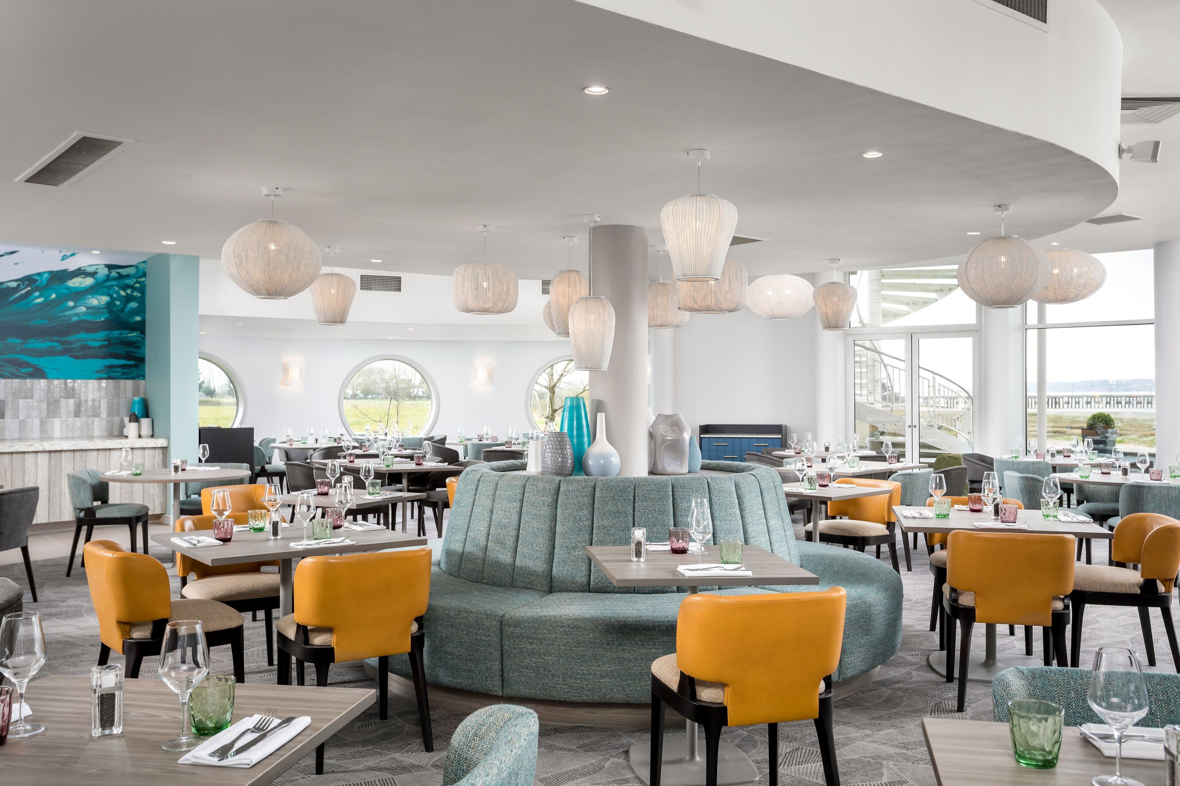 A modern restaurant with round tables, comfortable seating, and large windows offering a view.