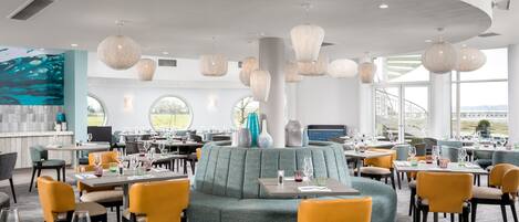 A modern restaurant with round tables, comfortable seating, and large windows offering a view.