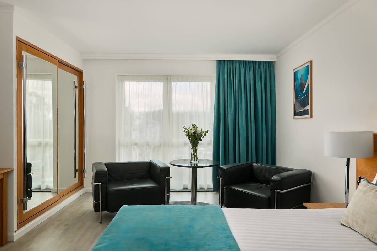A hotel room with a bed, two black leather chairs, a small round table with a vase of flowers, a large window with curtains, and a painting on the wall.