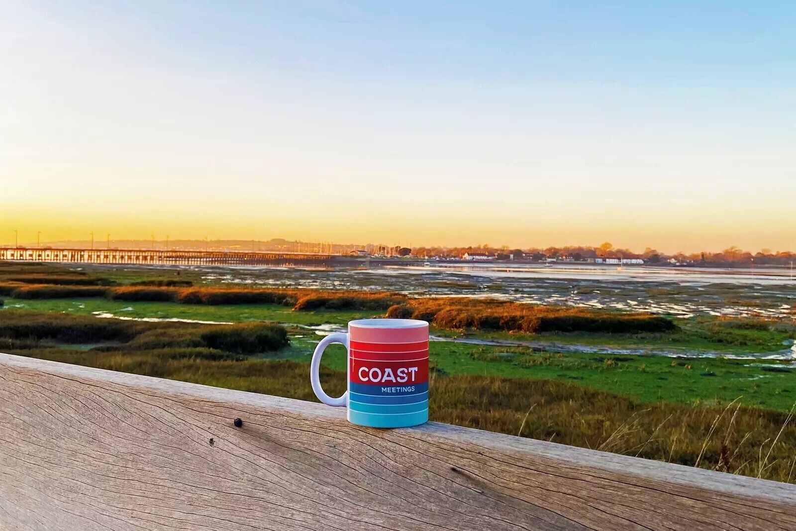 Eine Holzterrasse mit einer Tasse, auf der 'COAST MEETINGS' steht, und Blick auf eine Küstenlandschaft mit einem Pier in der Ferne.