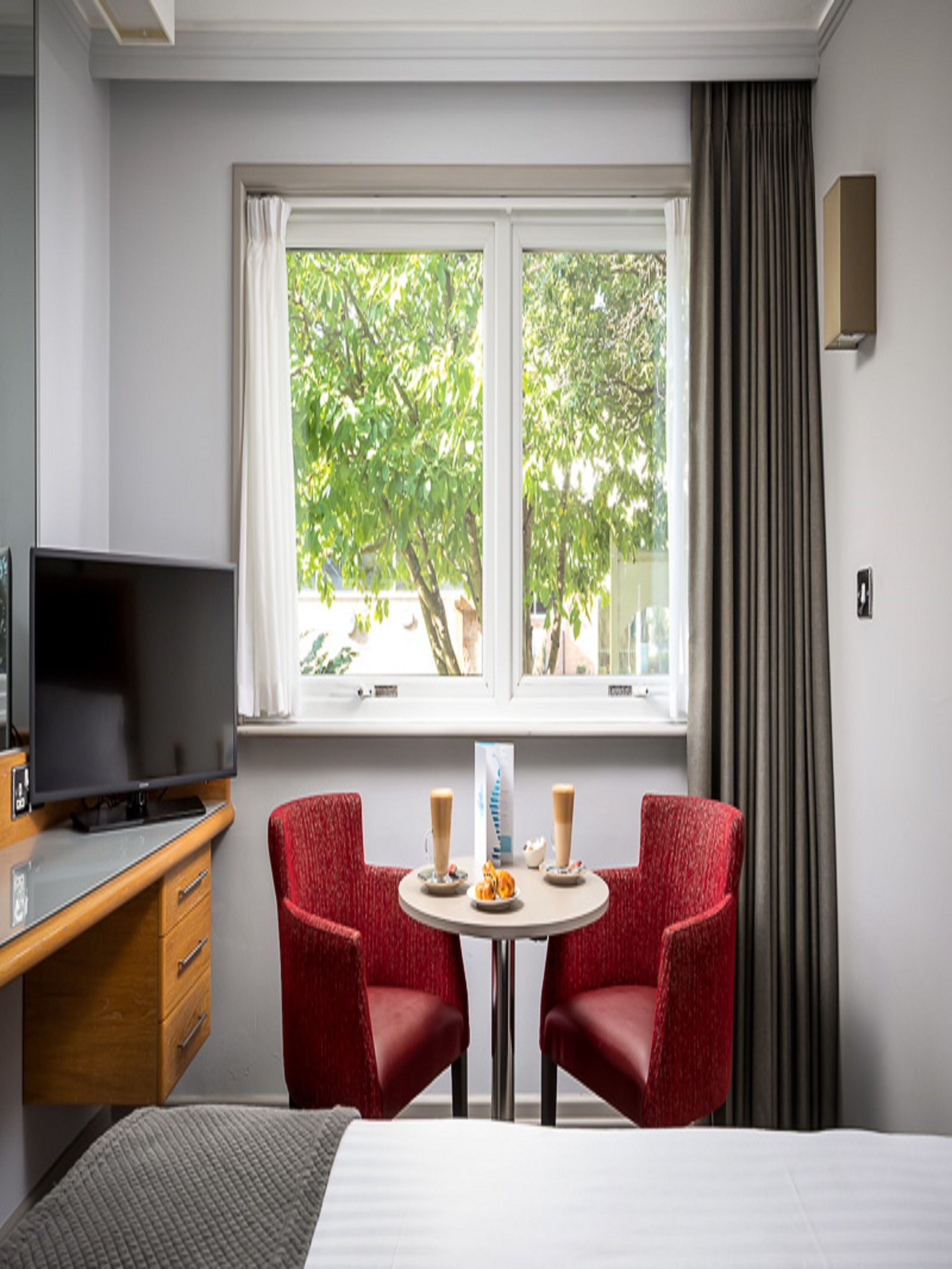 A hotel room with a bed, a desk with a TV, two red chairs around a small table with drinks, and a window with curtains.