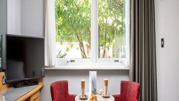 A hotel room with a bed, a desk with a TV, two red chairs around a small table with drinks, and a window with curtains.