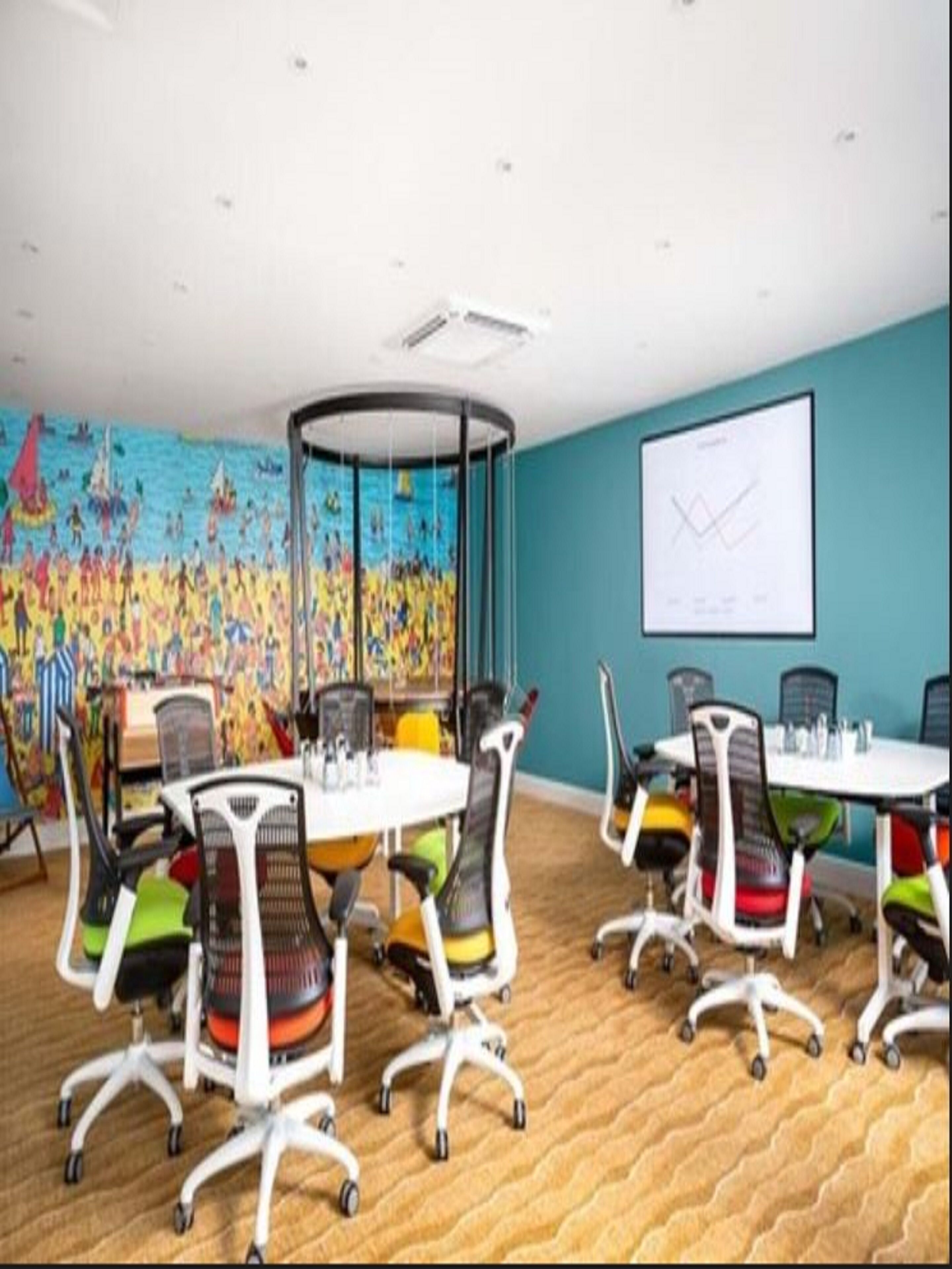 Un salón de conferencias con una mesa redonda, sillas coloridas, una pantalla de proyector y un mural vibrante en la pared.