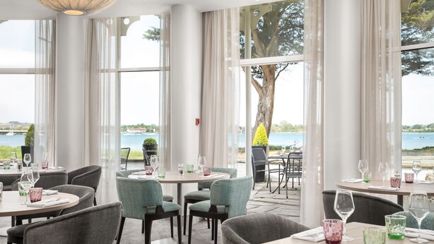 A restaurant with large windows offering a view of the sea and a tree. Tables are set with glasses and cutlery. - The Langstone Quays (Hayling Island)