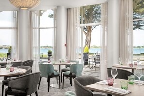 Un restaurant doté de grandes fenêtres donnant sur la mer et sur un arbre. Les tables sont dressées avec des verres et des couverts.