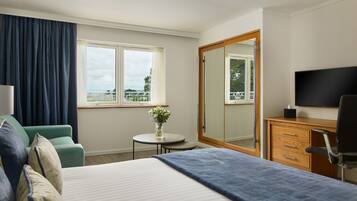 Quarto de hotel com cama, escrivaninha, cadeira, televisão, espelho, janela com cortinas e uma pequena mesa com um vaso de flores.