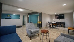 Eine Hotelhalle mit einer Rezeption, einem blauen Sofa, einem grauen Sessel, einem Holztisch und einem Essbereich mit Stühlen und einem an der Wand befestigten Fernseher.