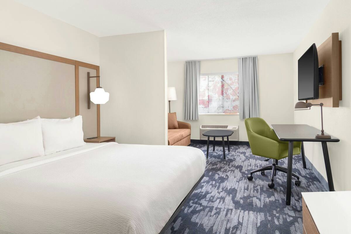A hotel room with a large bed, a desk, a green chair, a flat-screen TV mounted on the wall, and a window with curtains.