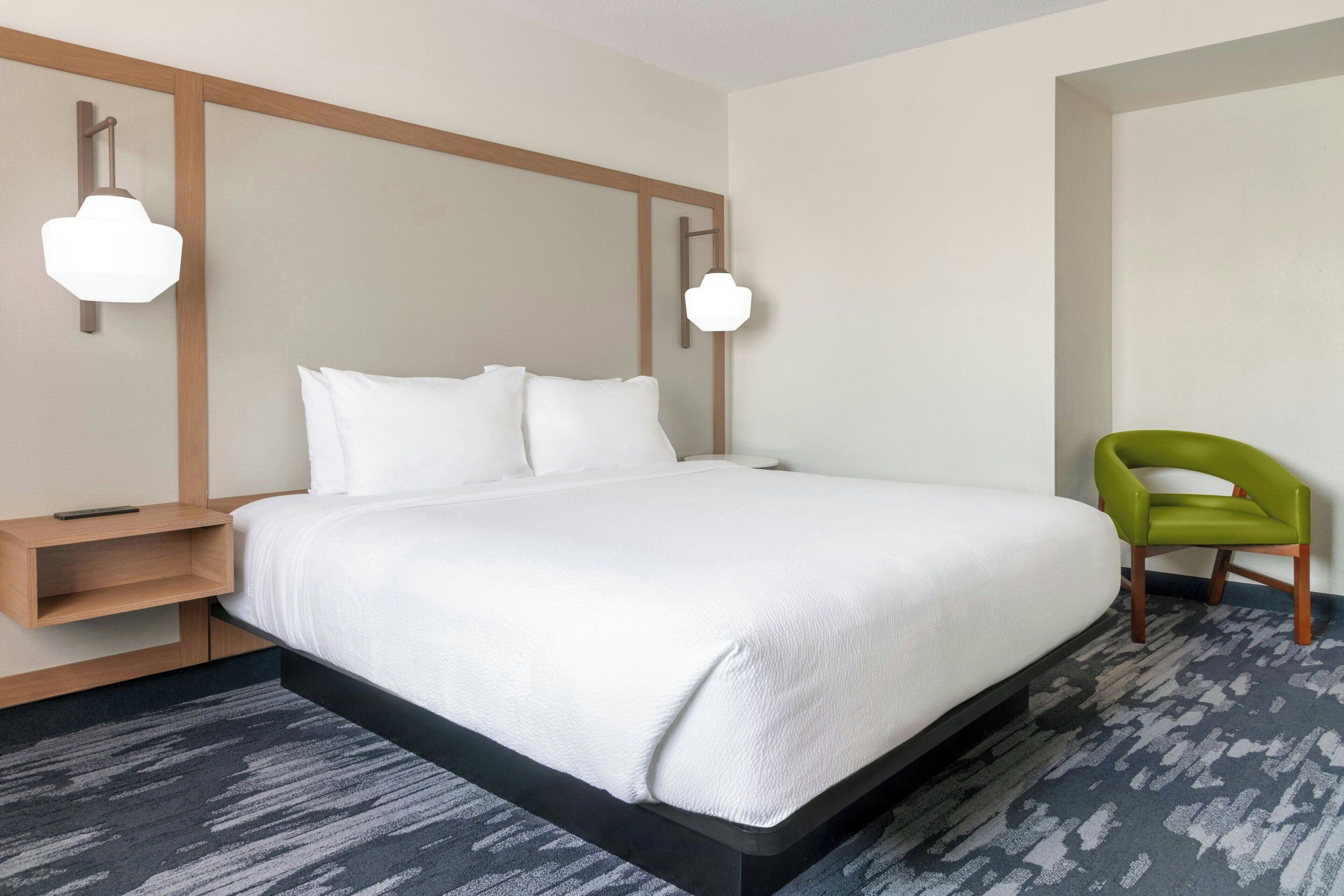 A modern hotel room with a large bed, a green chair, and a wooden nightstand.