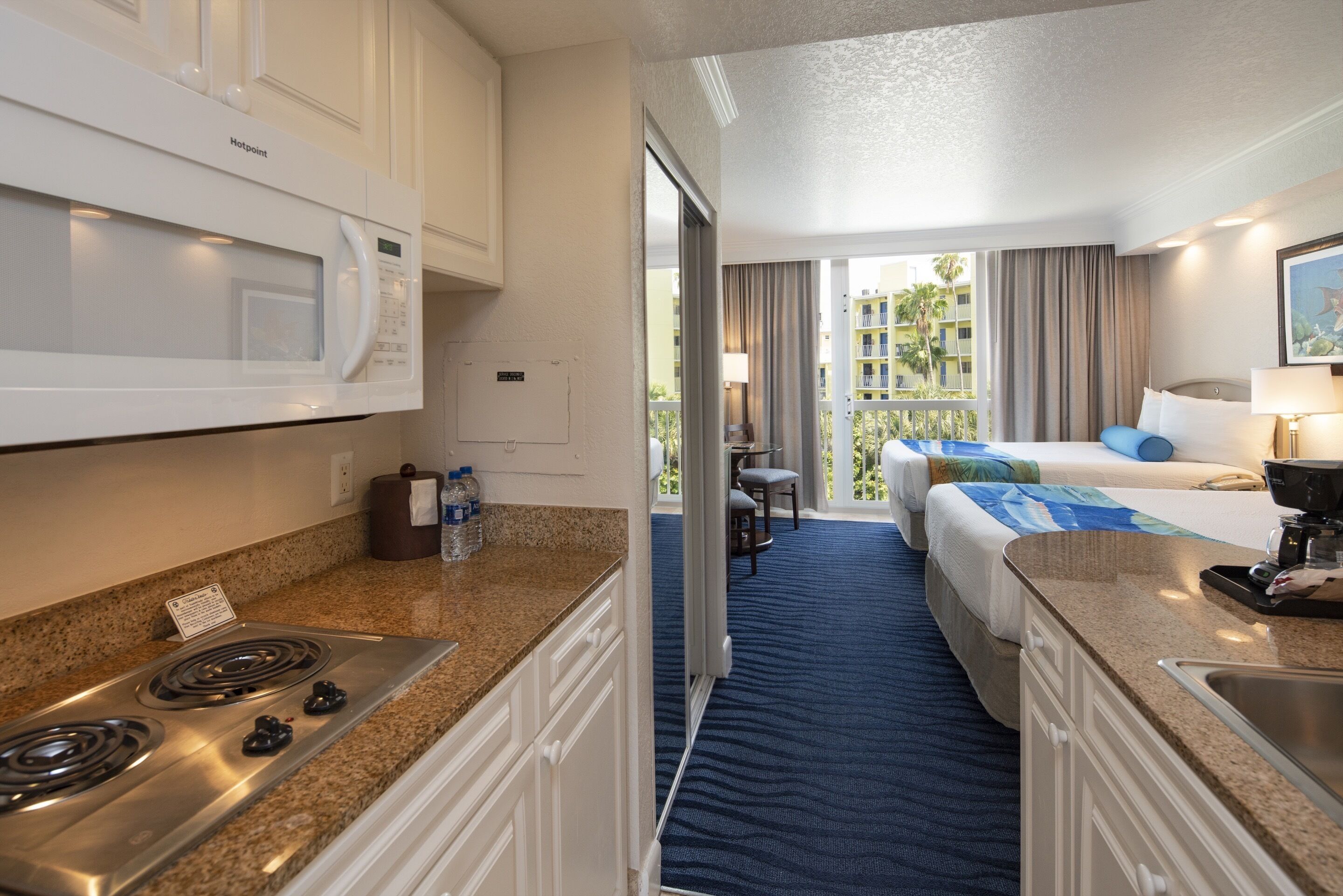A hotel room with a kitchenette, a bed with a blue and white bedspread, a balcony with a view of buildings, and a large window with curtains.