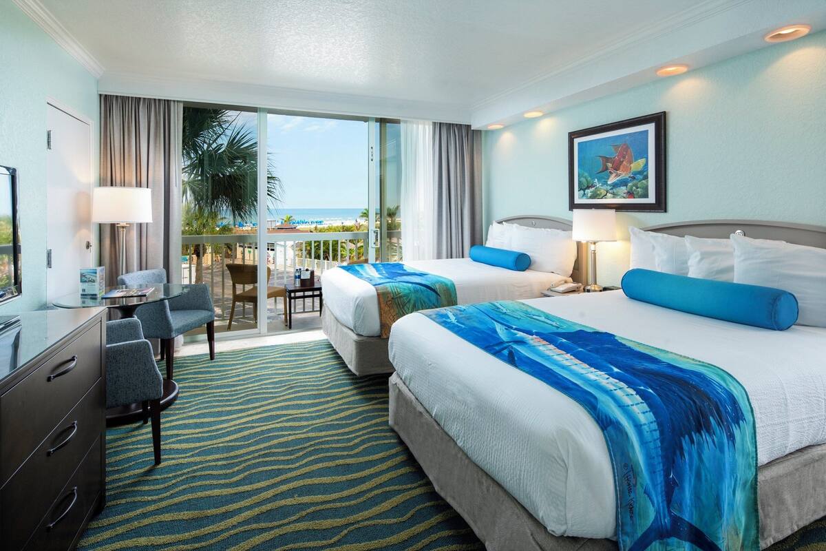 A hotel room with two beds, a desk, a chair, a balcony with a view of the beach, and a painting on the wall.