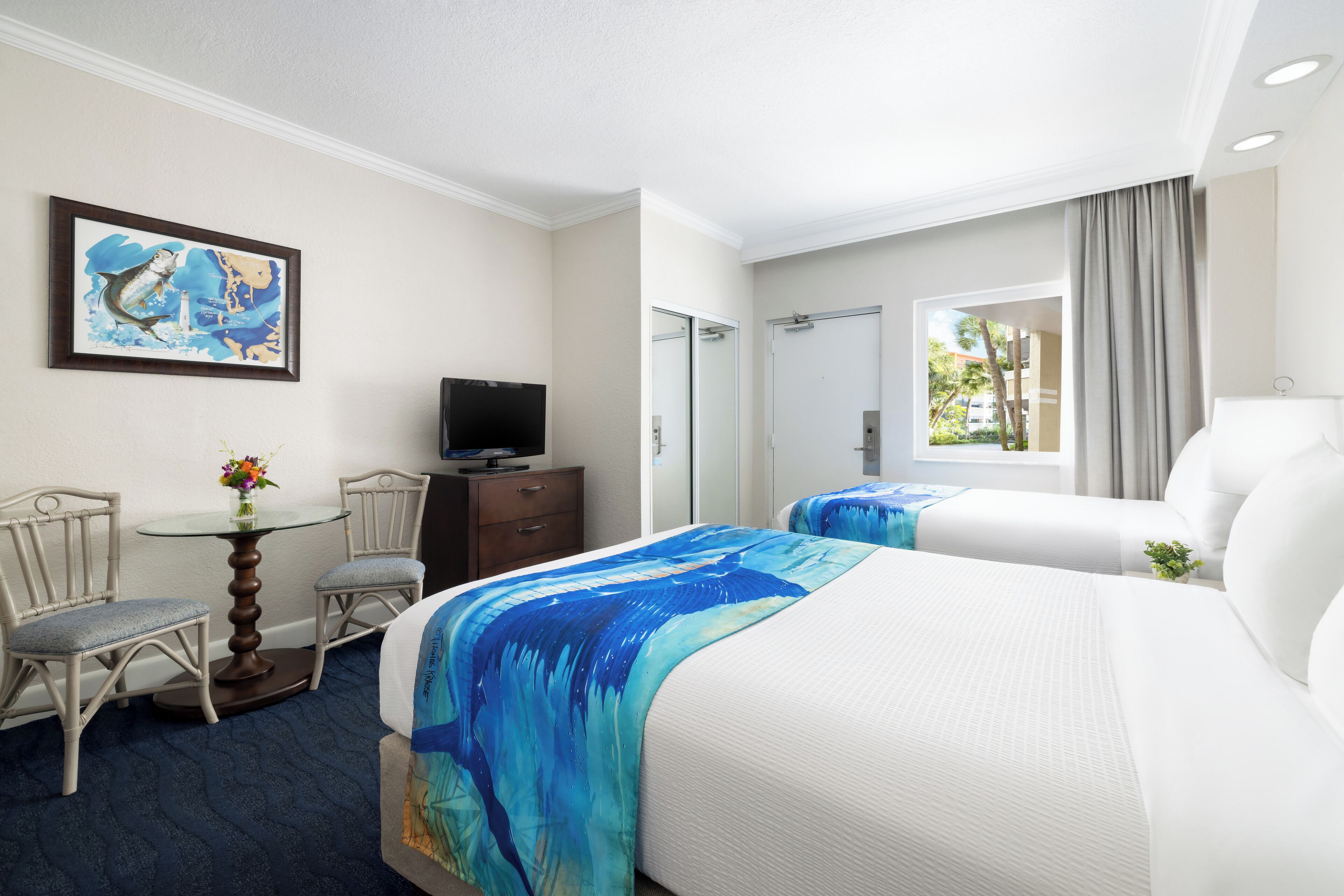 Habitación de hotel con dos camas, un televisor, un escritorio con una silla, una mesita con un florero y un cuadro en la pared.